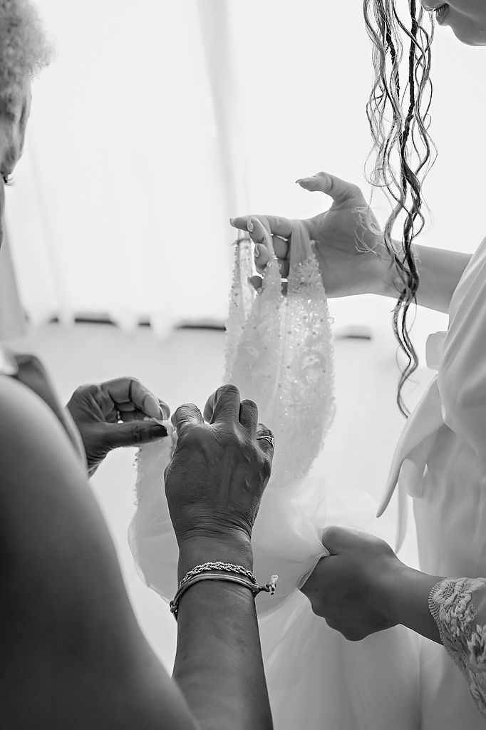 Several pairs of hands delicately adjust a white lace wedding dress in a candid black and white photograph.