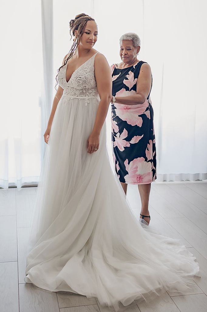 Two women admire a flowing white wedding dress in a bright, sunlit room with white decor.