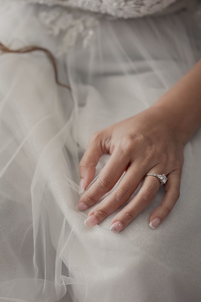 An elegant engagement ring sparkles on a hand resting delicately against white tulle fabric.