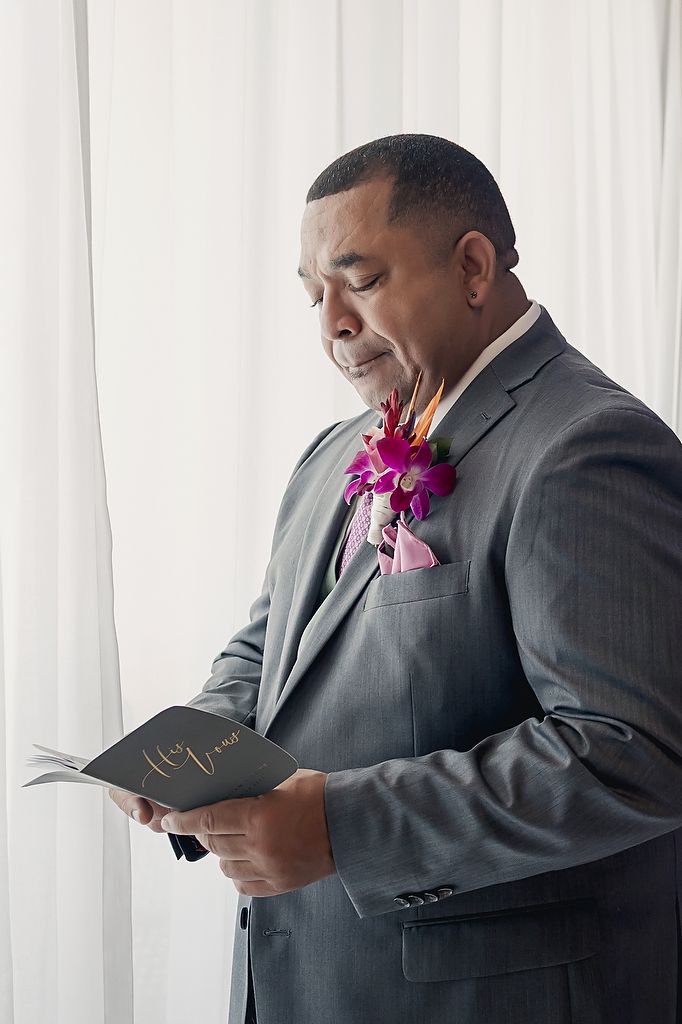 A man in a grey suit with a pink orchid boutonniere reads from a small black book beside white curtains.