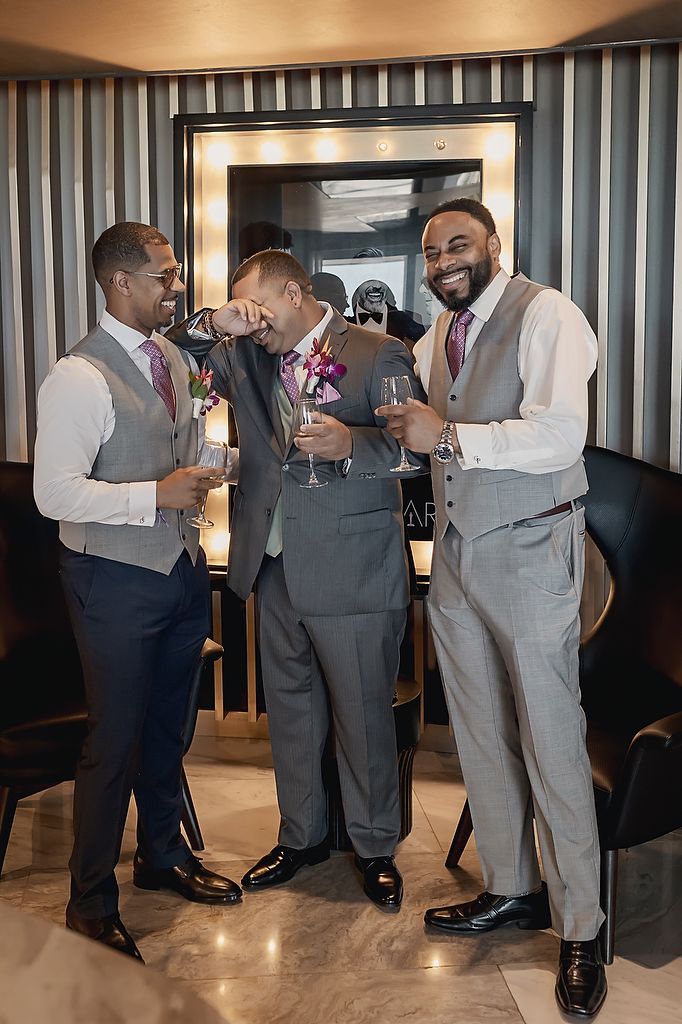 Three men in formal gray vests and suits share laughter and drinks at an elegant indoor venue with modern decor.