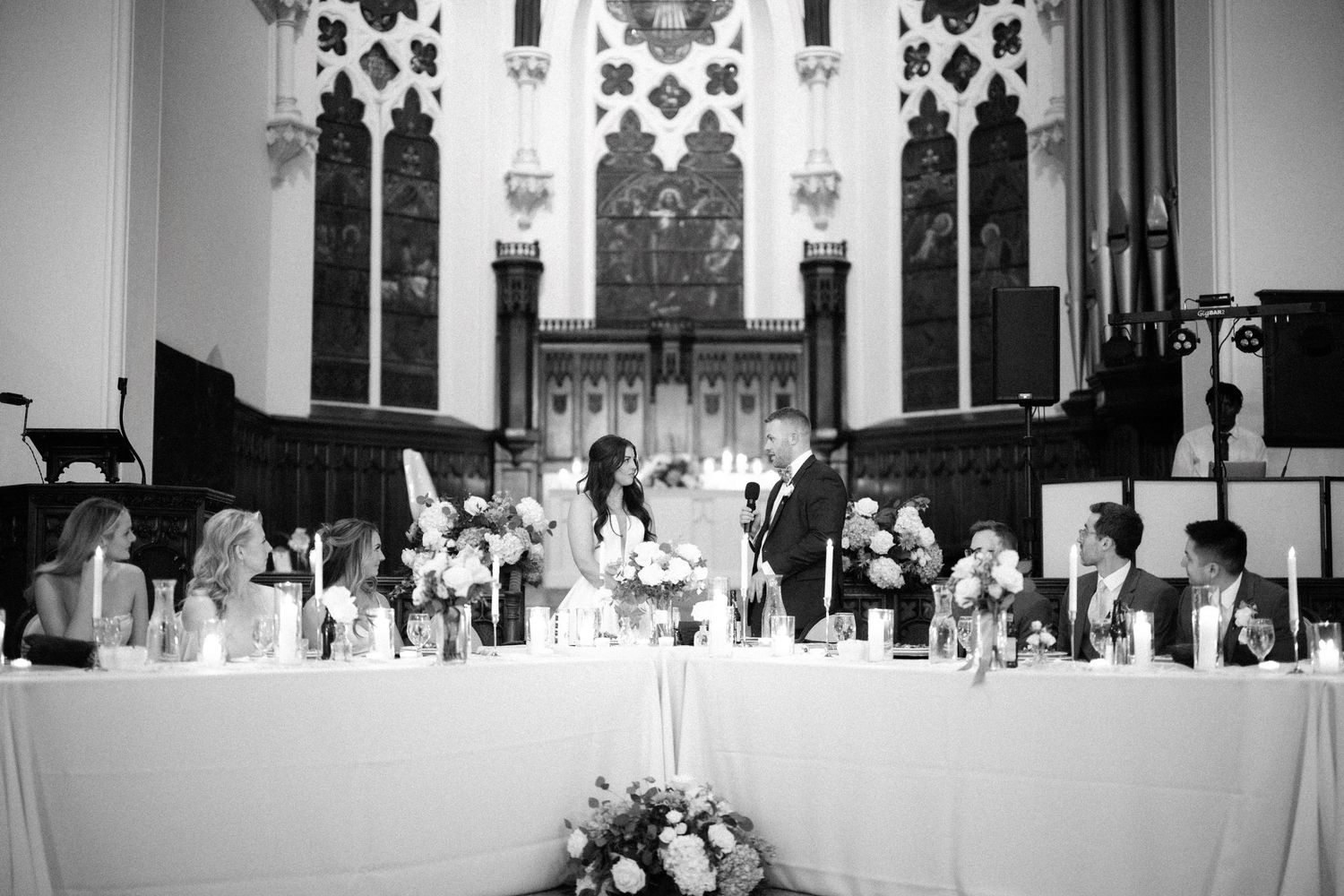 Black and white photo of a formal wedding reception head table with elaborate church architecture and floral decor.