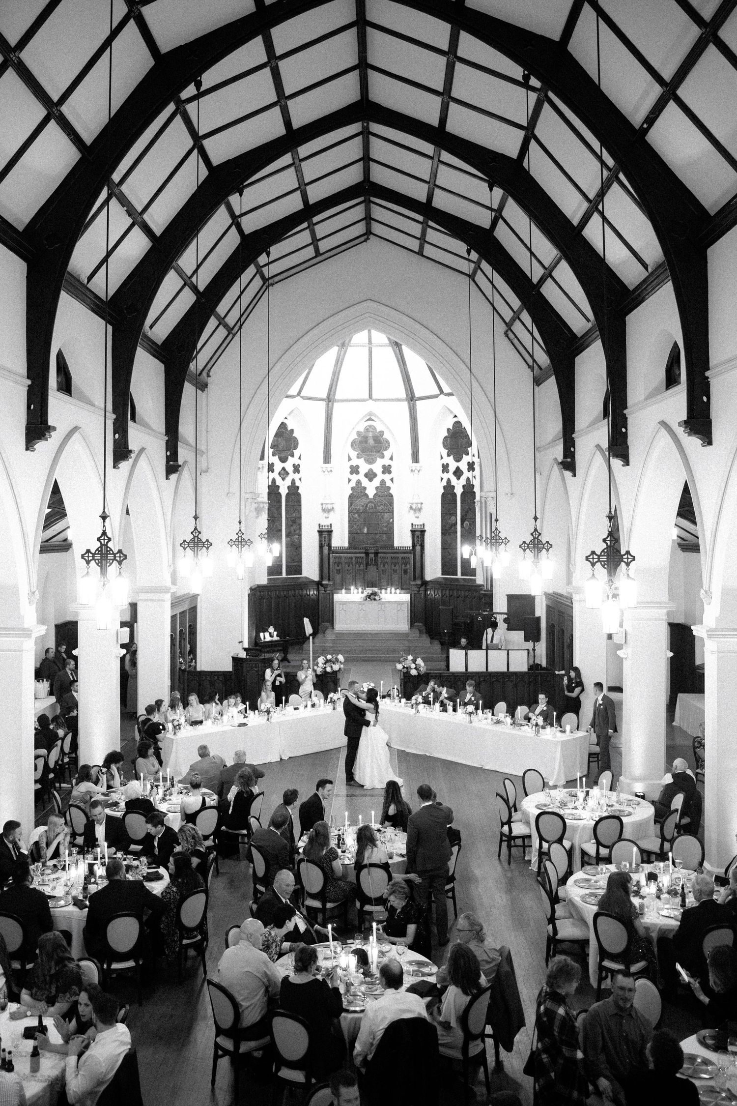 A black and white photograph of a wedding reception in a grand Gothic-style church hall with vaulted ceilings and stained glass windows.
