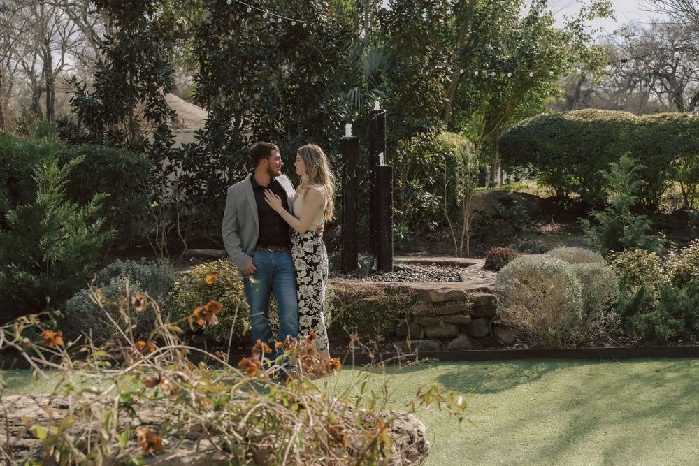 Two people embrace while standing in a lush garden landscape with colorful flowers and greenery on a sunny day.