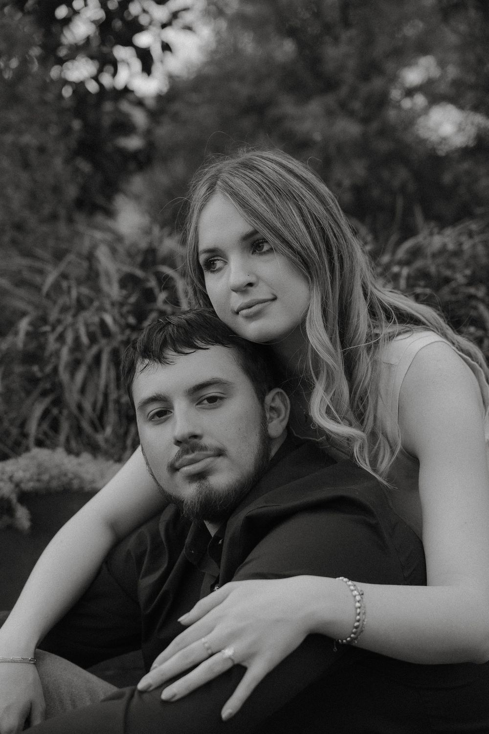 An intimate black and white portrait of a couple embracing outdoors with natural foliage in the background.