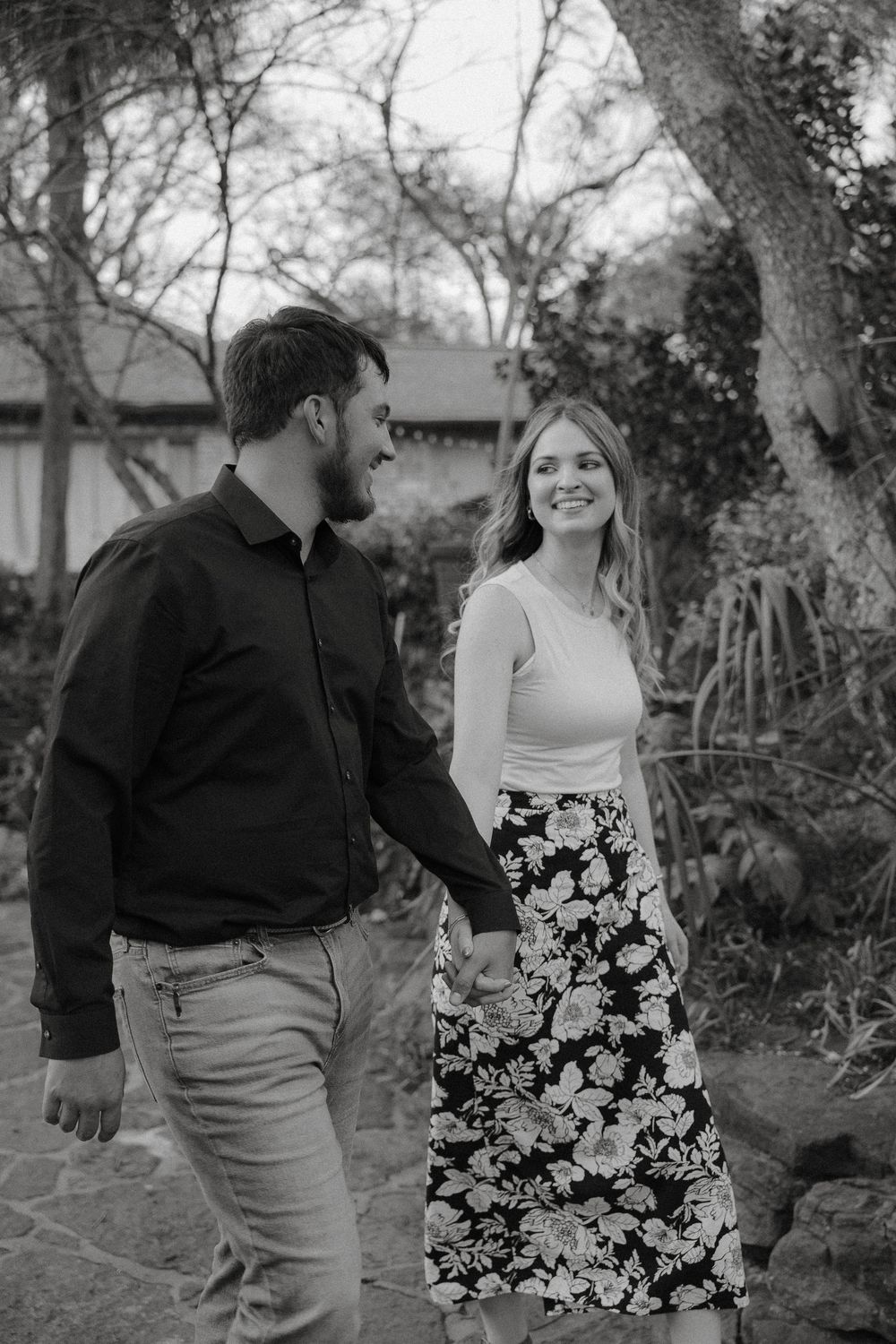 A romantic black and white photo of a couple walking together in a garden, with her wearing a floral skirt.