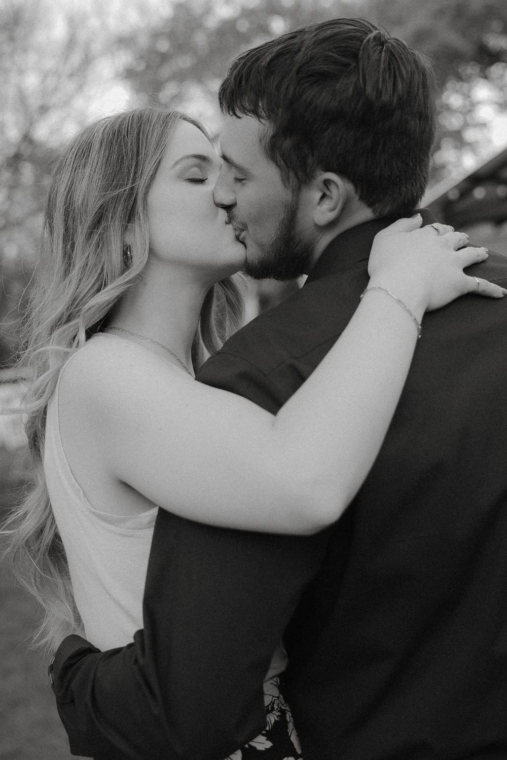 A romantic black and white portrait shows a couple sharing an intimate kiss outdoors.