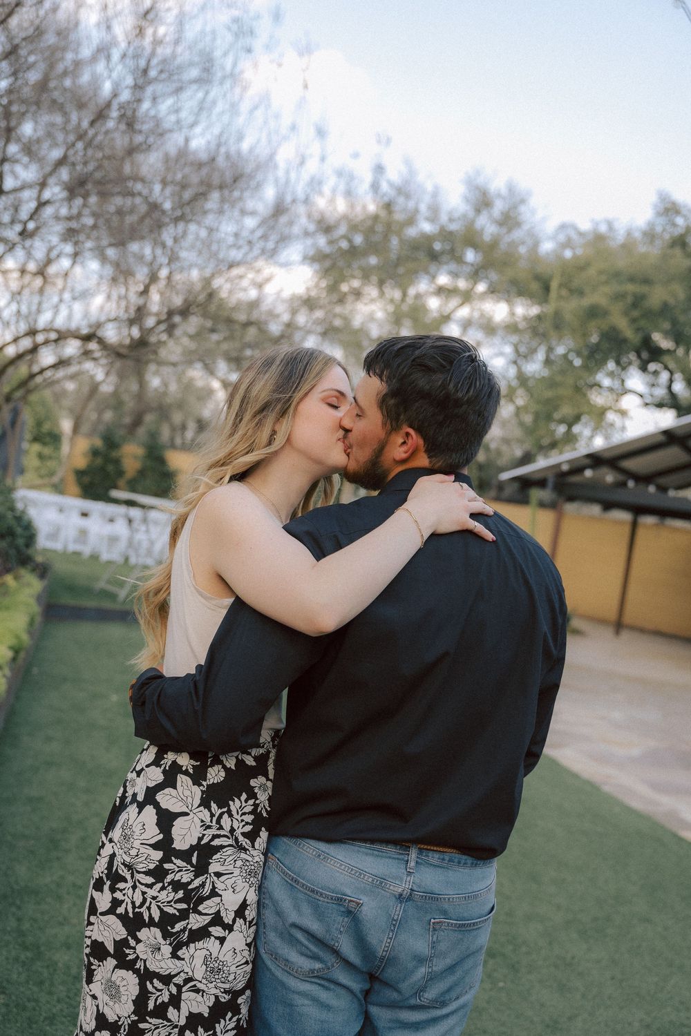 A couple embraces romantically in an outdoor garden setting with a floral dress and casual attire.