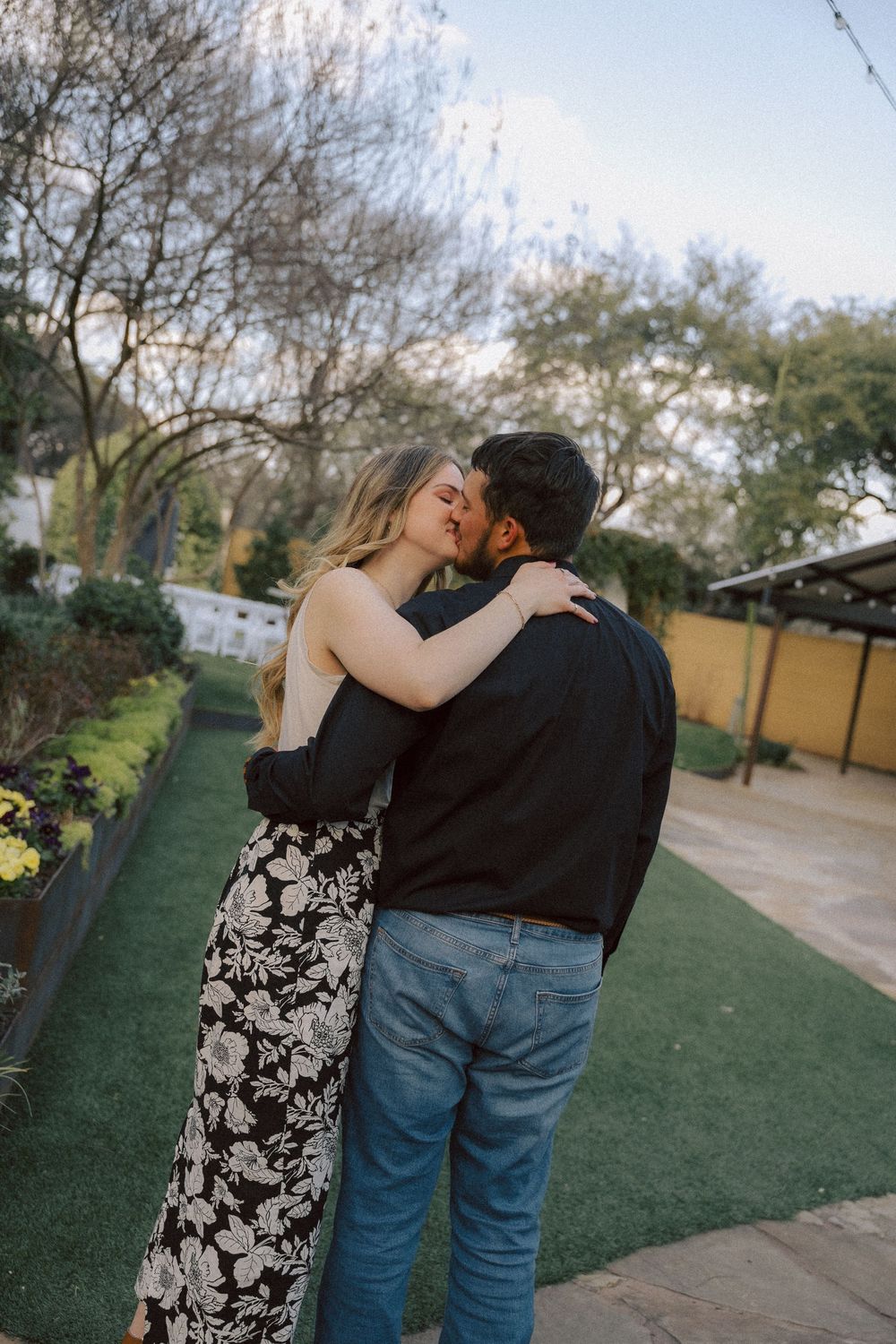 A couple embraces on an outdoor green walkway surrounded by trees and a yellow wall during sunset.