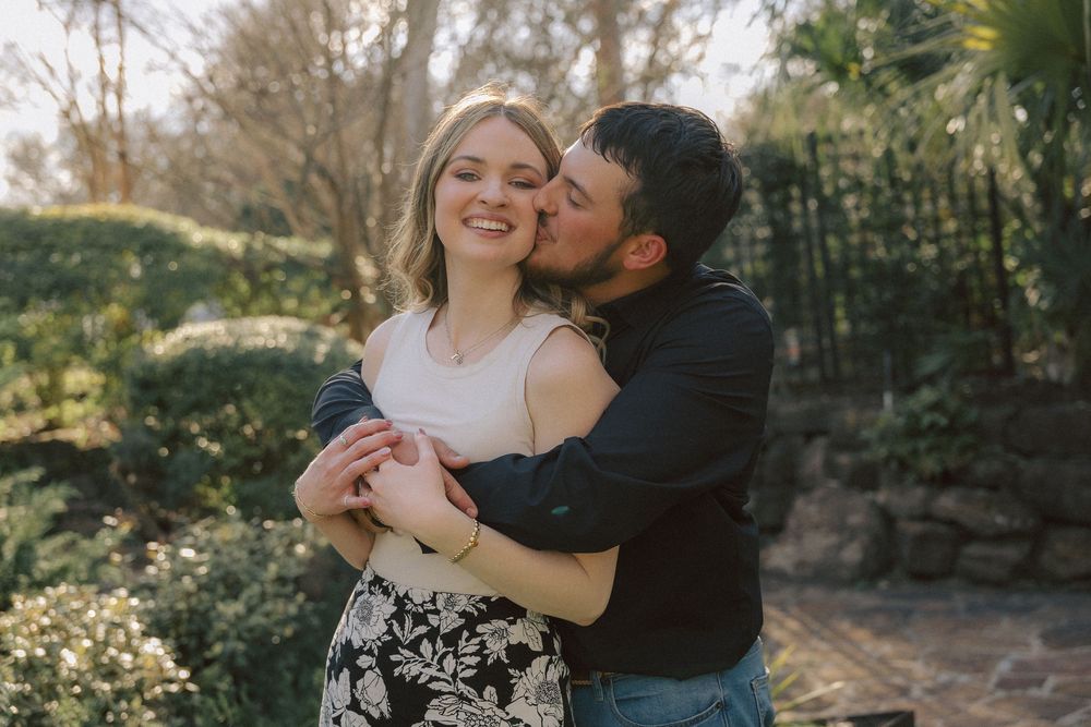 A couple embraces in a sunny garden setting with floral landscaping and natural lighting.