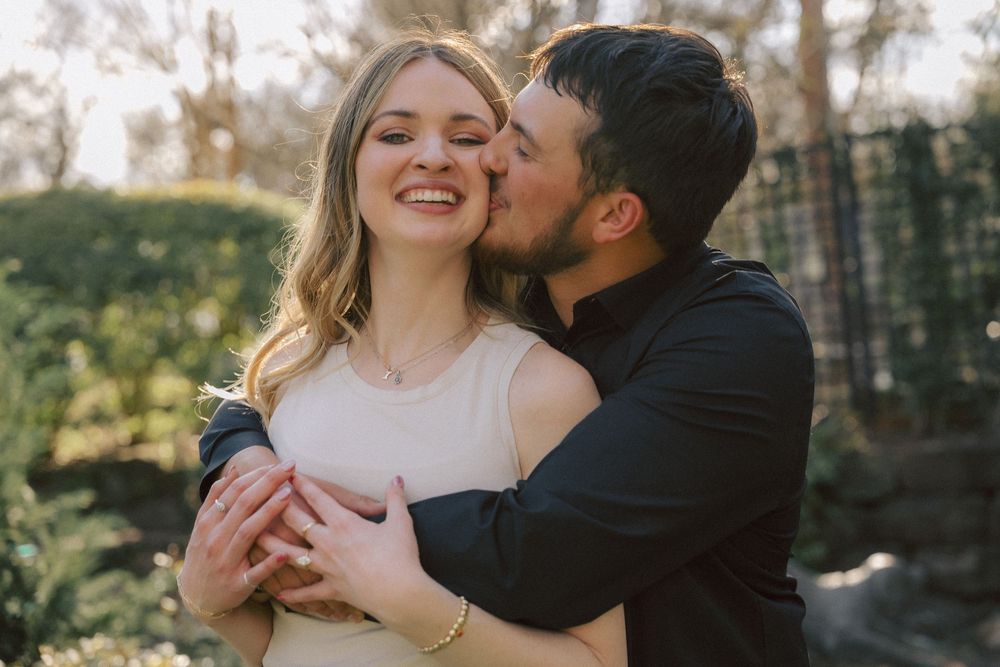 An affectionate couple embraces outdoors on a sunny day with trees in the background.