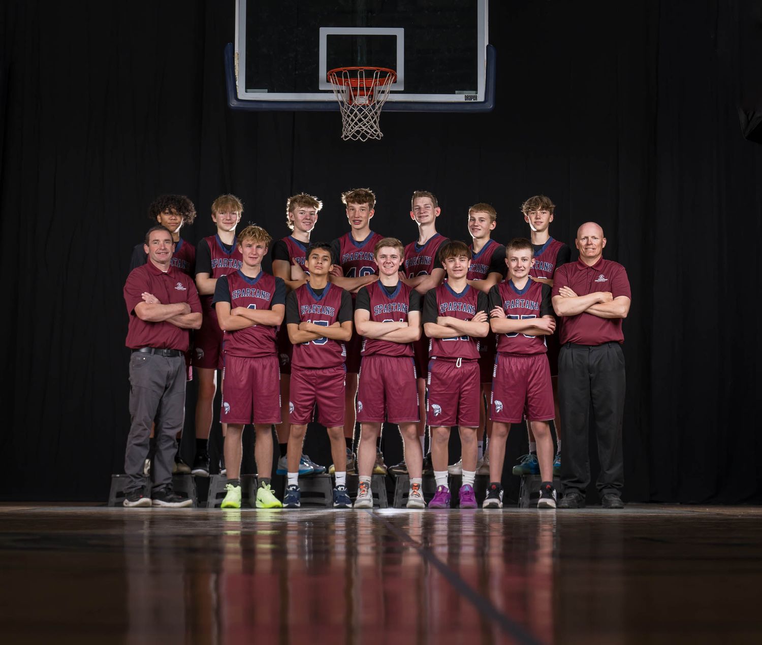 Youth basketball team in maroon uniforms poses for team photo on indoor court with coaches on either end.