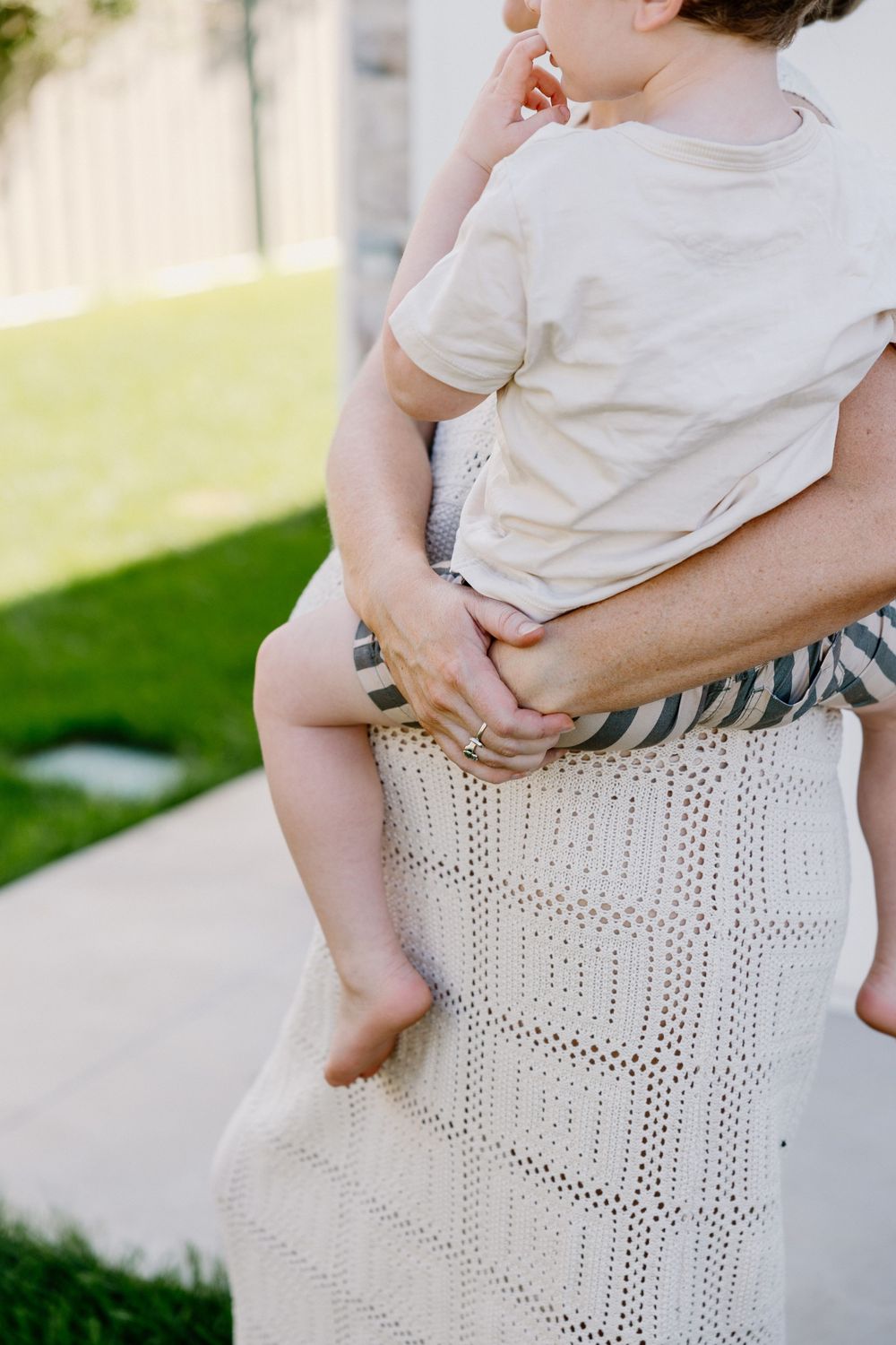 Child wearing white t-shirt being held in arms against a cream colored crochet dress in outdoor sunlight setting.