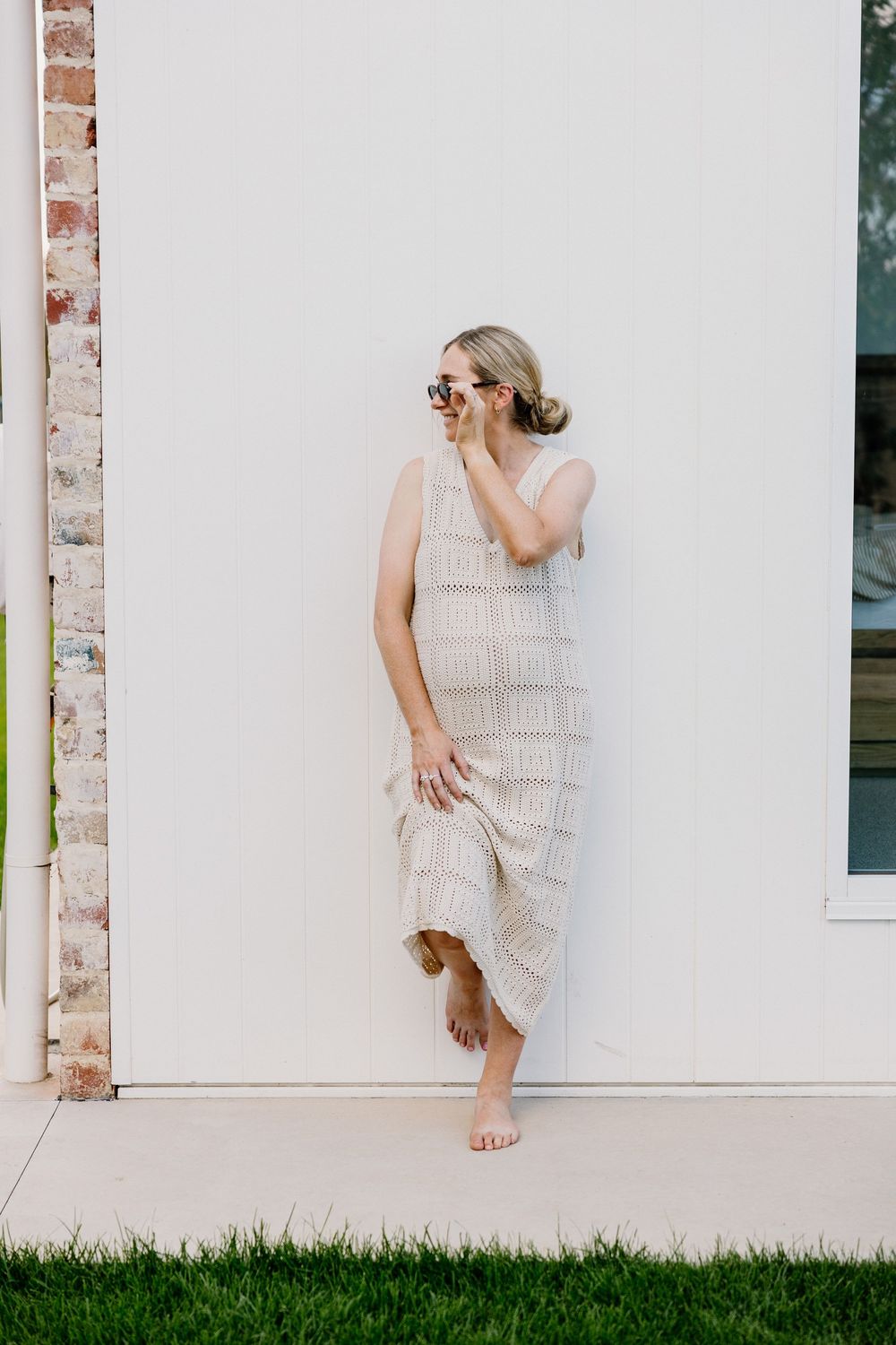 A figure in a flowing white lace dress poses playfully against a white wall with grass below.