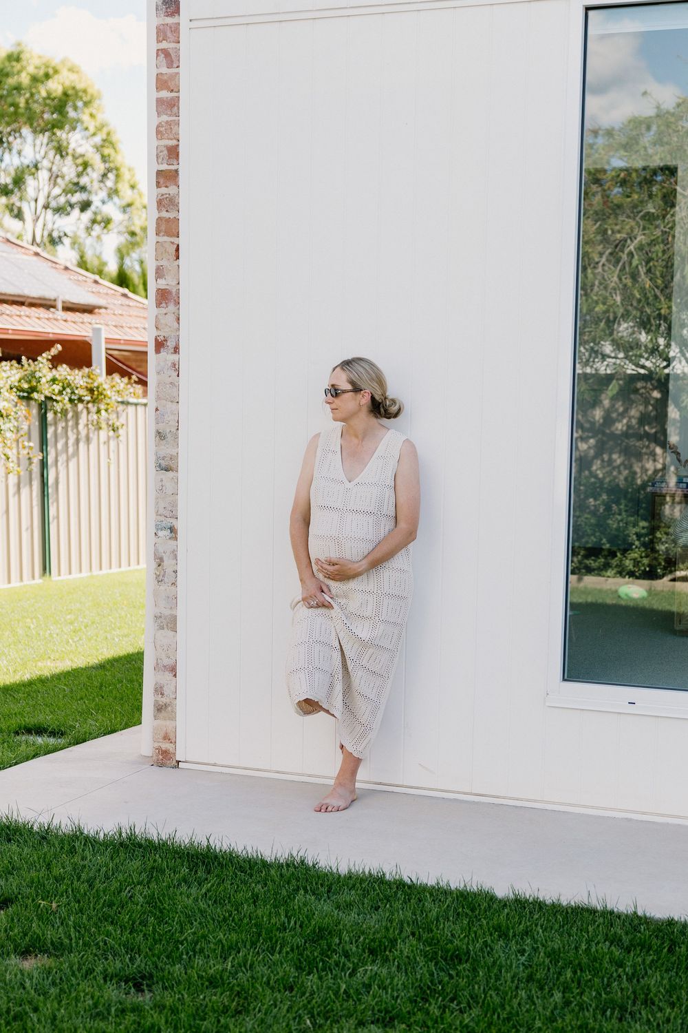 A person in a light linen dress leans against a white exterior wall of a modern home with manicured lawn.