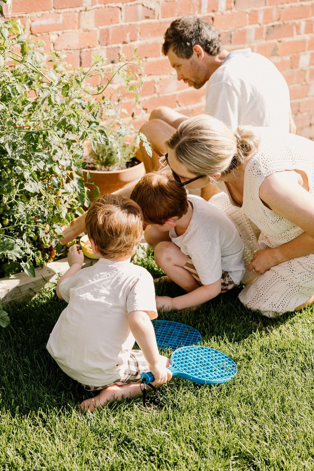 Group gathered around a blue garden kneeler pad examining plants near a brick wall in sunlit backyard garden.