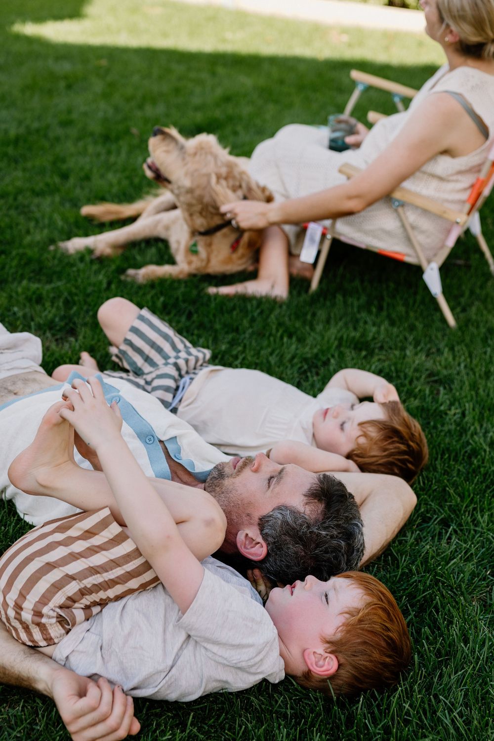 Family relaxing together on a sunny lawn with a dog lounging in a chair nearby.