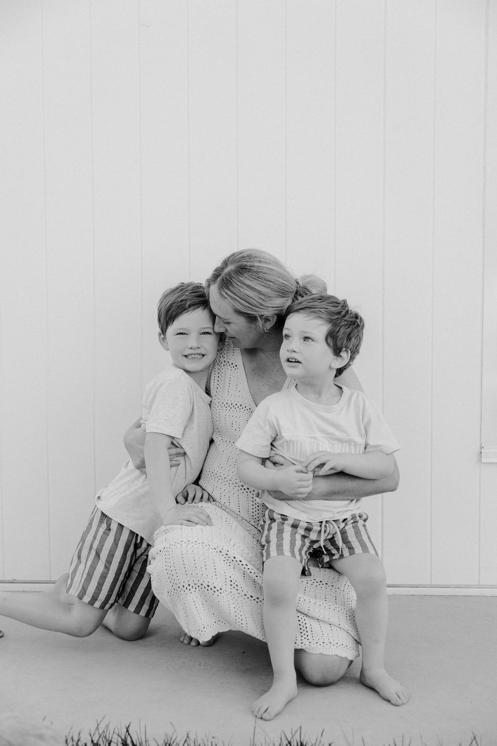 A heartwarming black and white portrait of a parent embracing two young children wearing matching striped shorts.