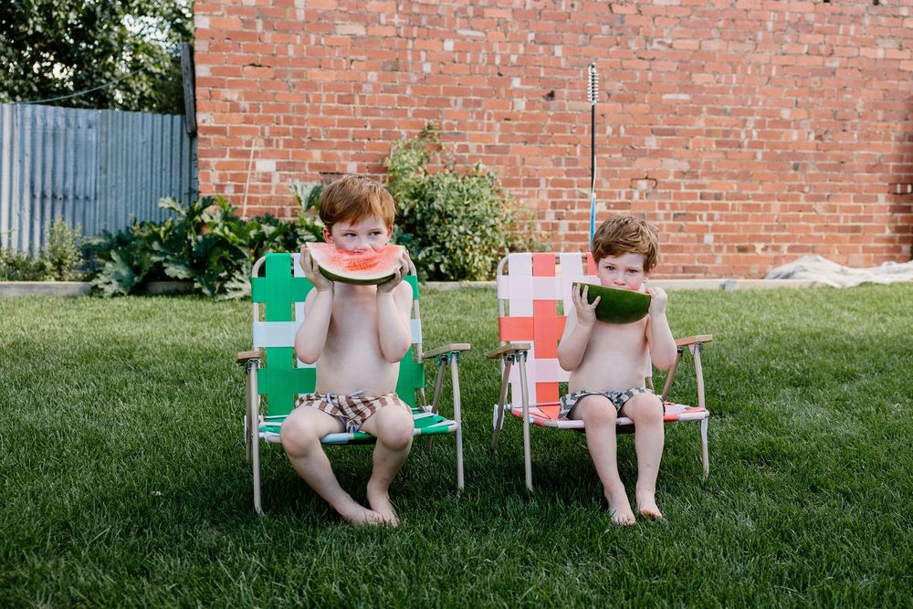 Two young children sitting in colorful lawn chairs on grass enjoying slices of watermelon on a summer day.