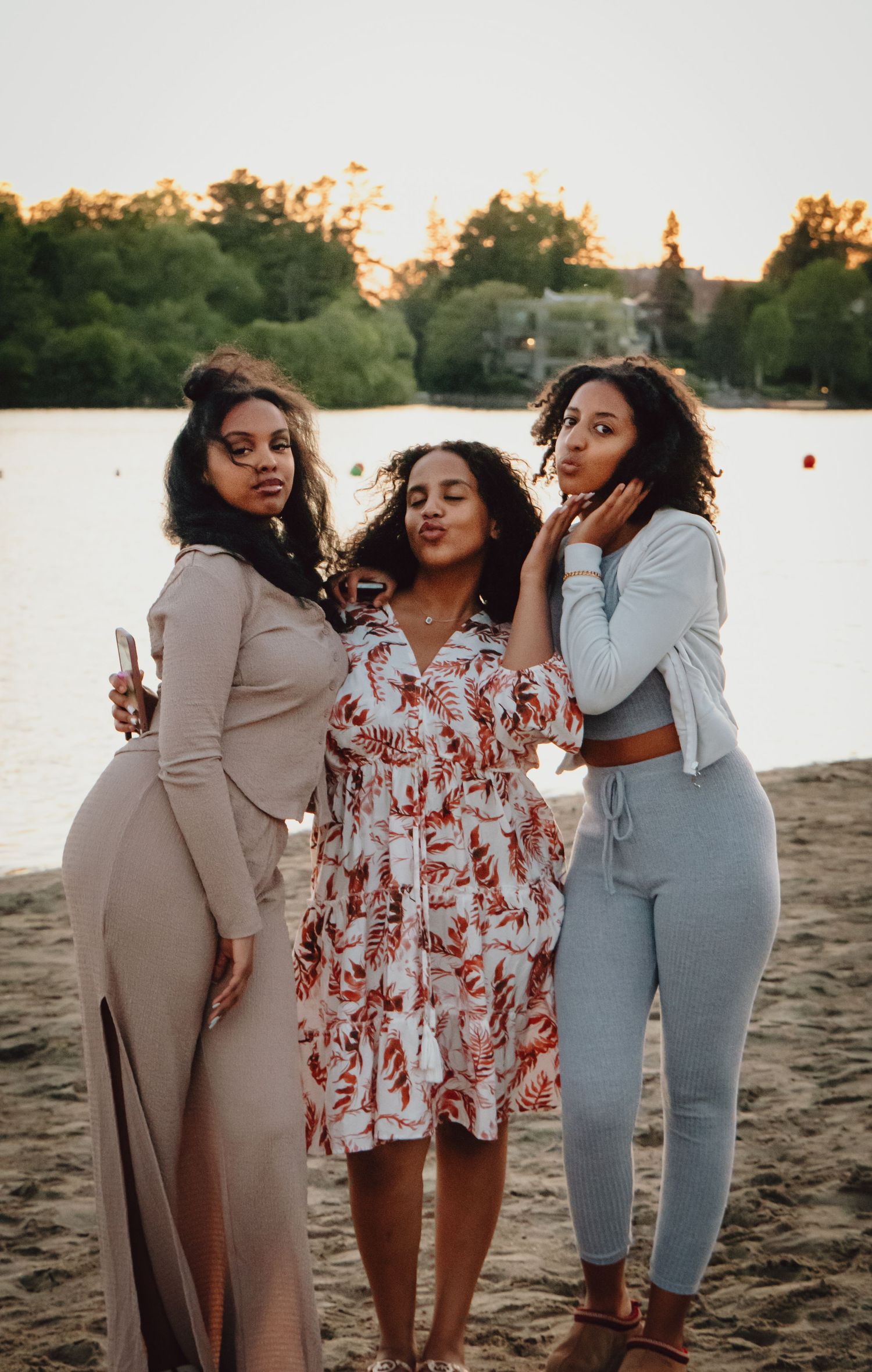 Three friends pose playfully on a beach at sunset with a lake in the background.