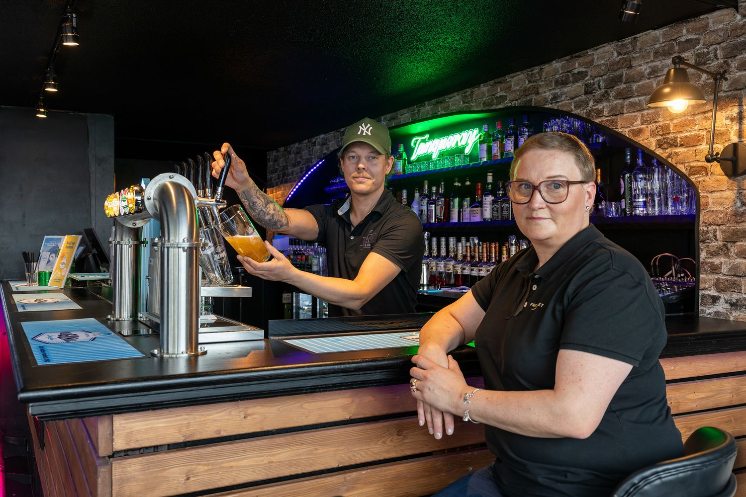 Two bar staff members serving drinks at a modern bar with neon lights and bottle display shelves.