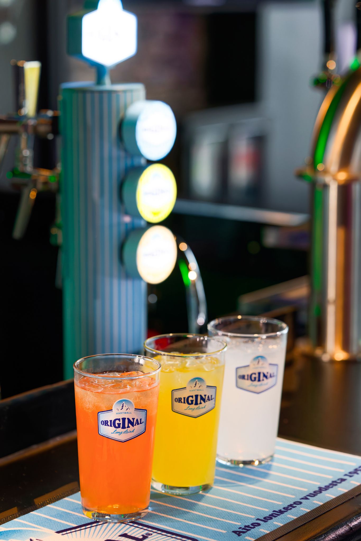 Three colorful Original brand drinks in glasses on a bar counter with beer taps in the background.