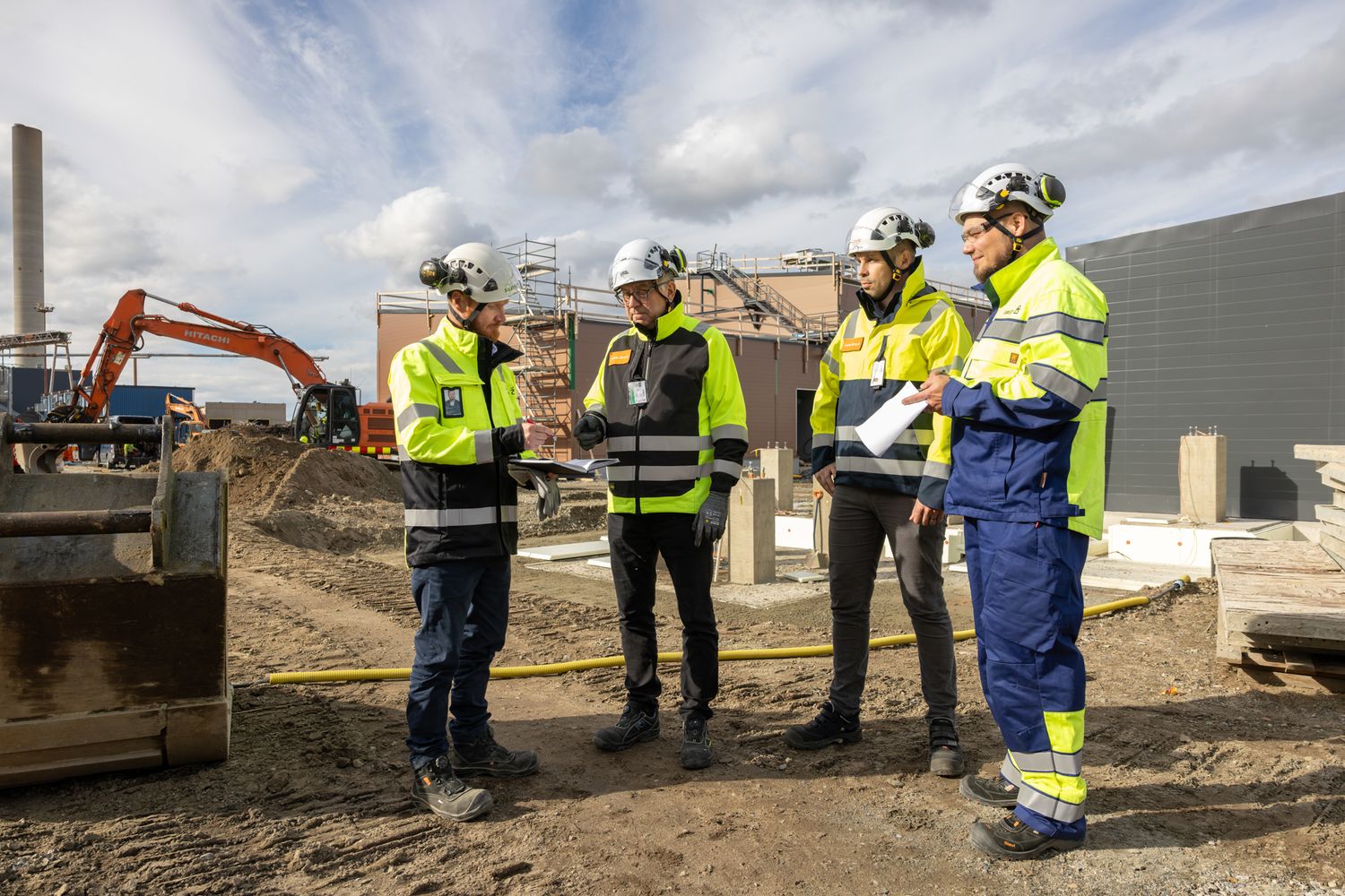 Construction workers in safety gear gather at an outdoor worksite with excavation equipment and machinery.
