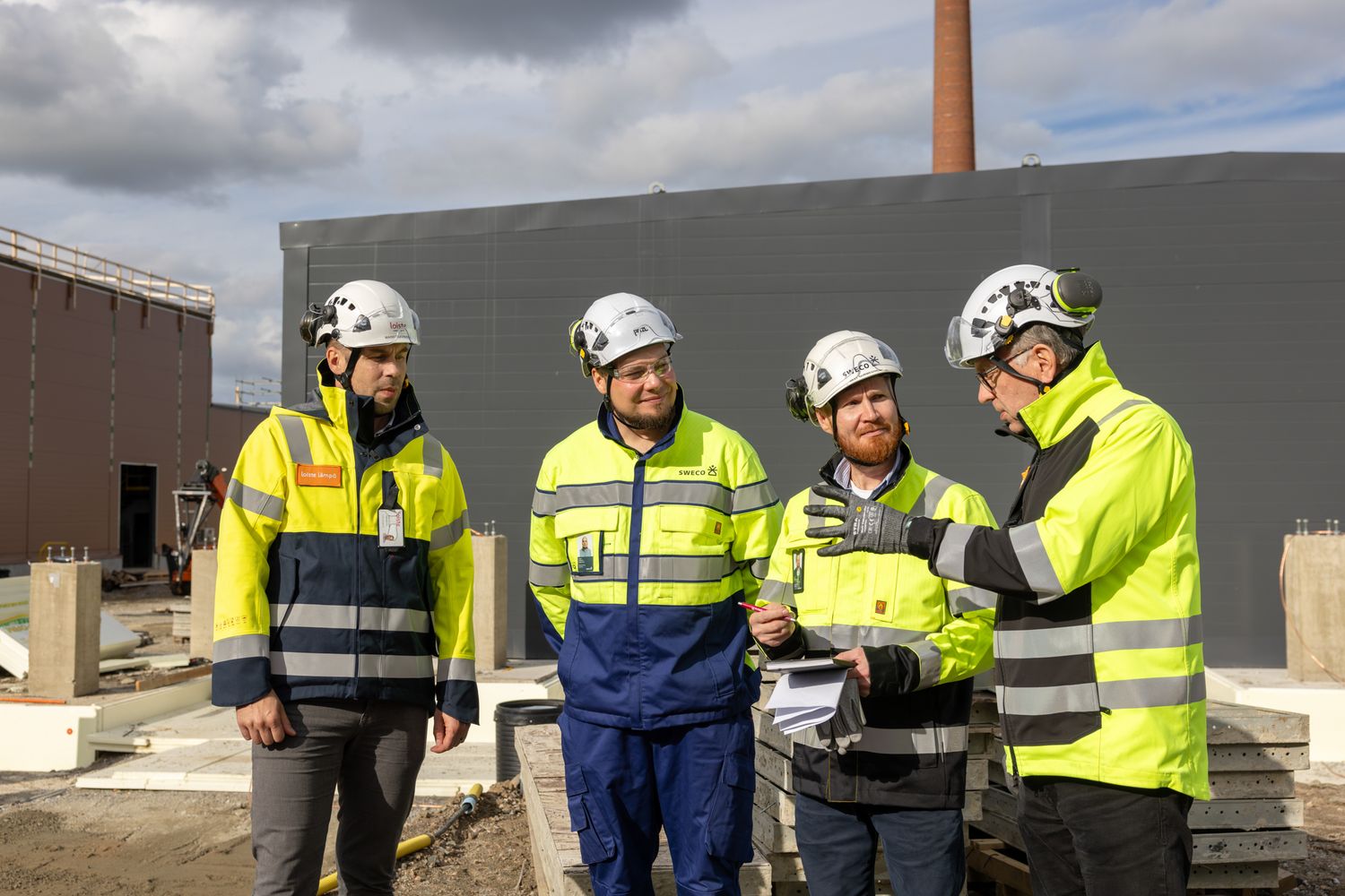 Workers wearing high-visibility vests and hard hats collaborate at a construction site with industrial buildings.