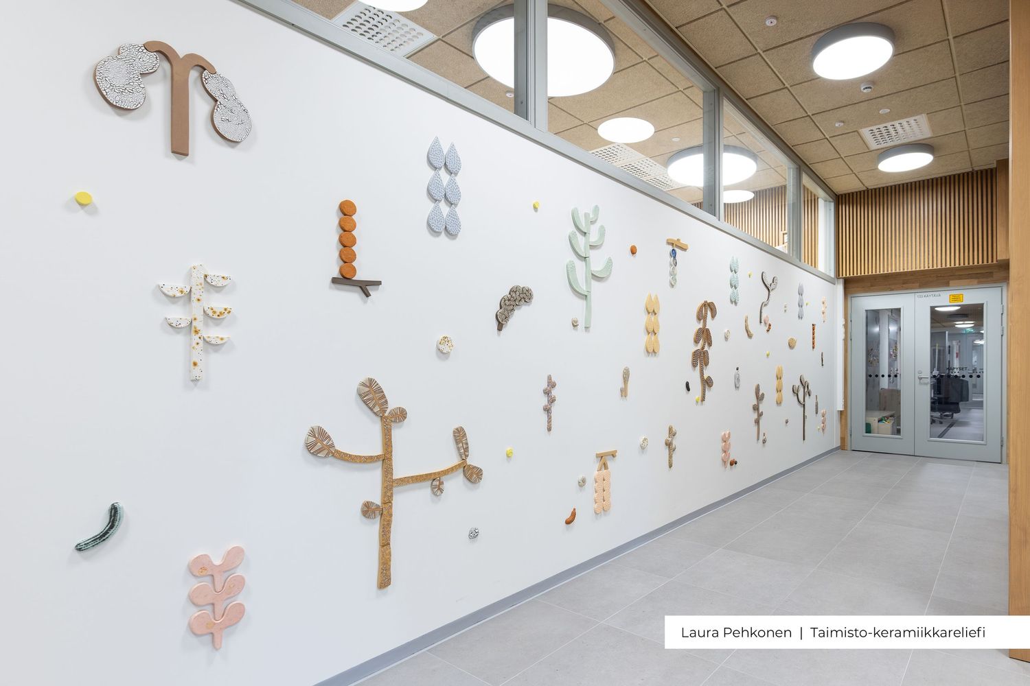 Colorful ceramic art installation on a white wall in a modern hallway, featuring abstract nature-inspired shapes and forms.