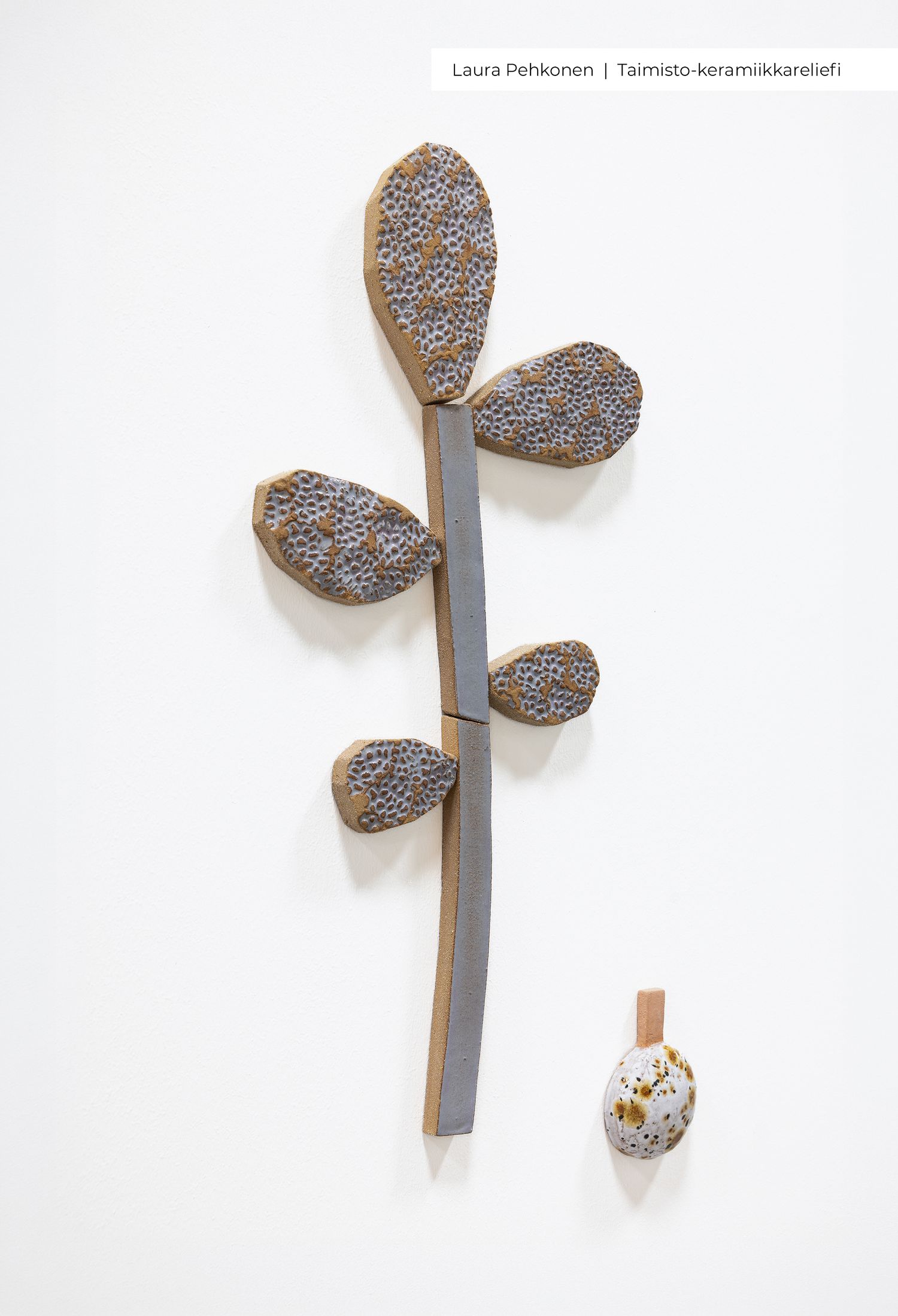 Ceramic wall relief sculpture by Laura Pehkonen featuring leaf-shaped textured pieces on a stem, displayed on a white gallery wall.