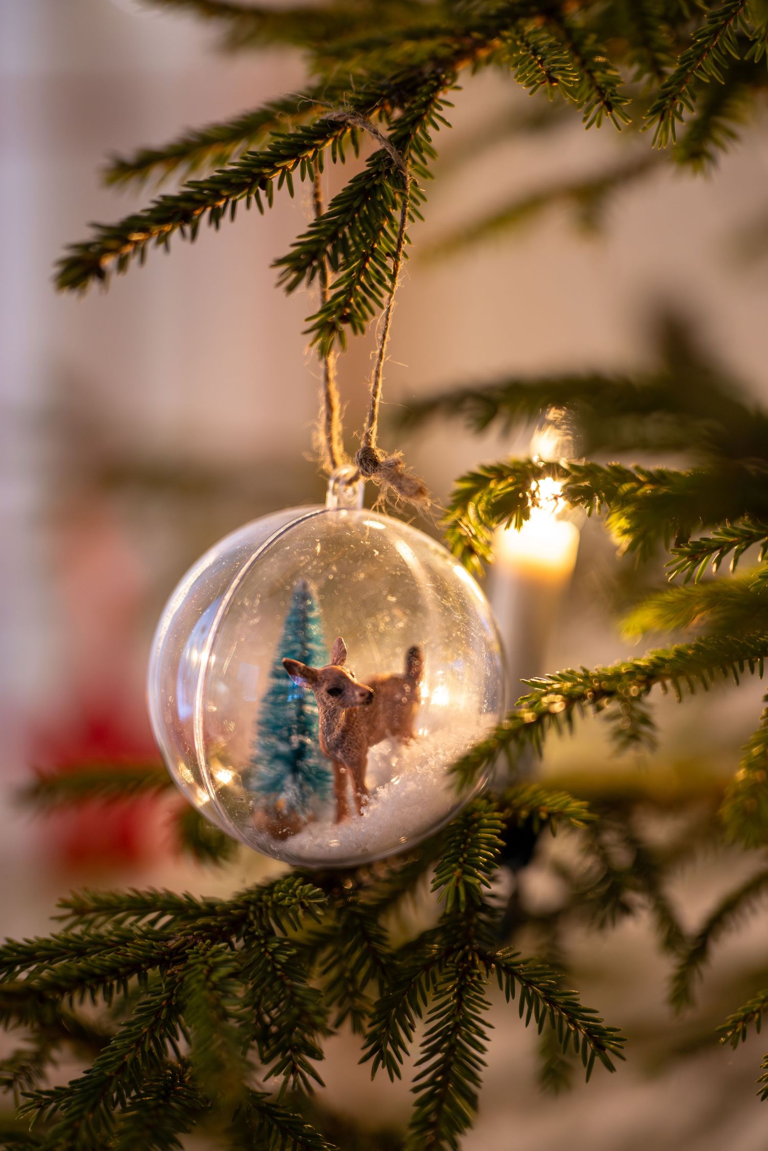 Clear glass Christmas ornament with miniature winter scene hanging on evergreen tree branch illuminated by warm light.