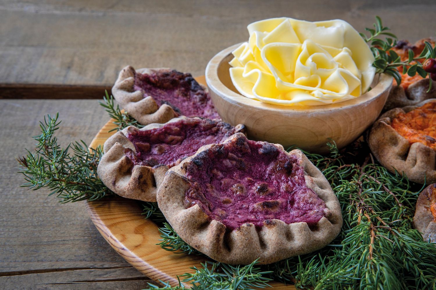 Traditional Finnish Karelian pies with purple filling on wooden board, served with butter roses in bowl.