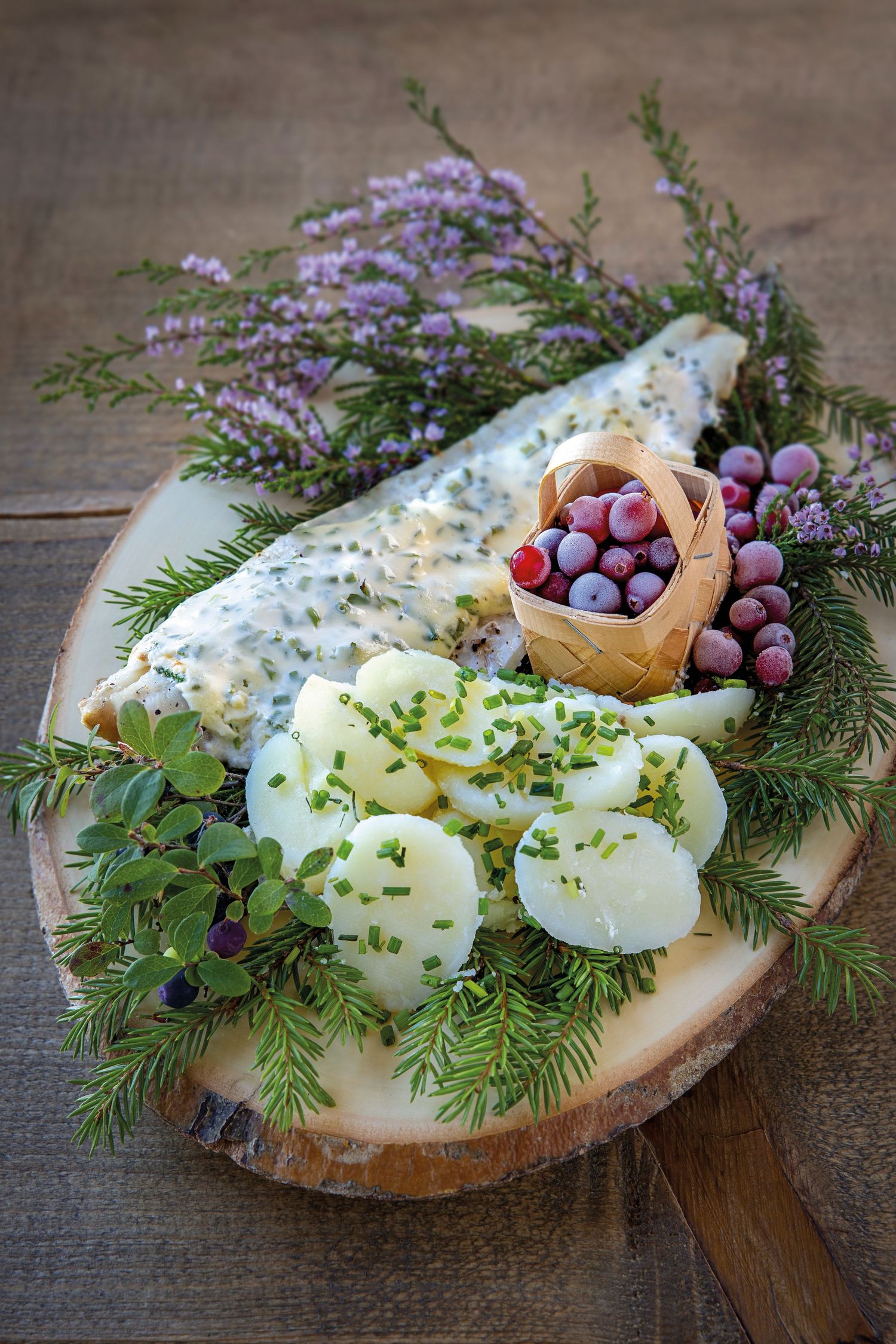 Smoked fish fillet on rustic wooden board with potato salad, fresh herbs, berries in birch basket, and heather garnish.