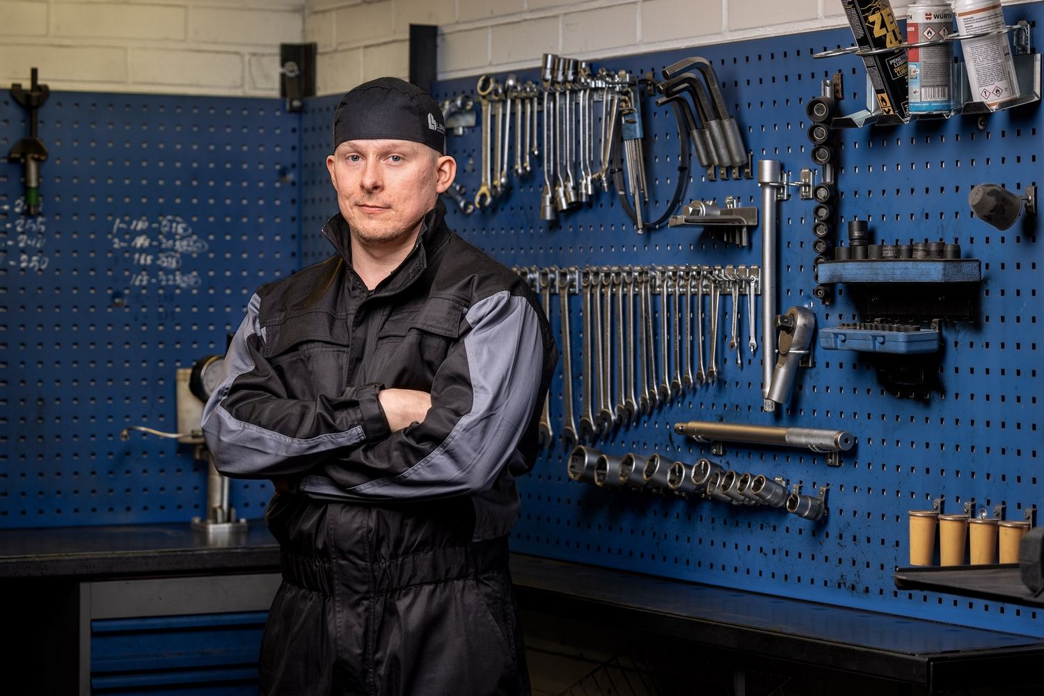 Male mechanic in black uniform stands confidently with crossed arms in front of organized tool wall in professional garage.