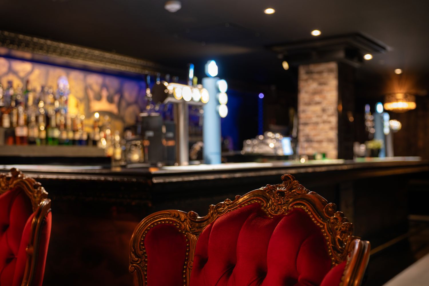 Elegant bar interior with red velvet gold-framed chairs, dark countertop, beer taps, and softly lit shelves of bottles.
