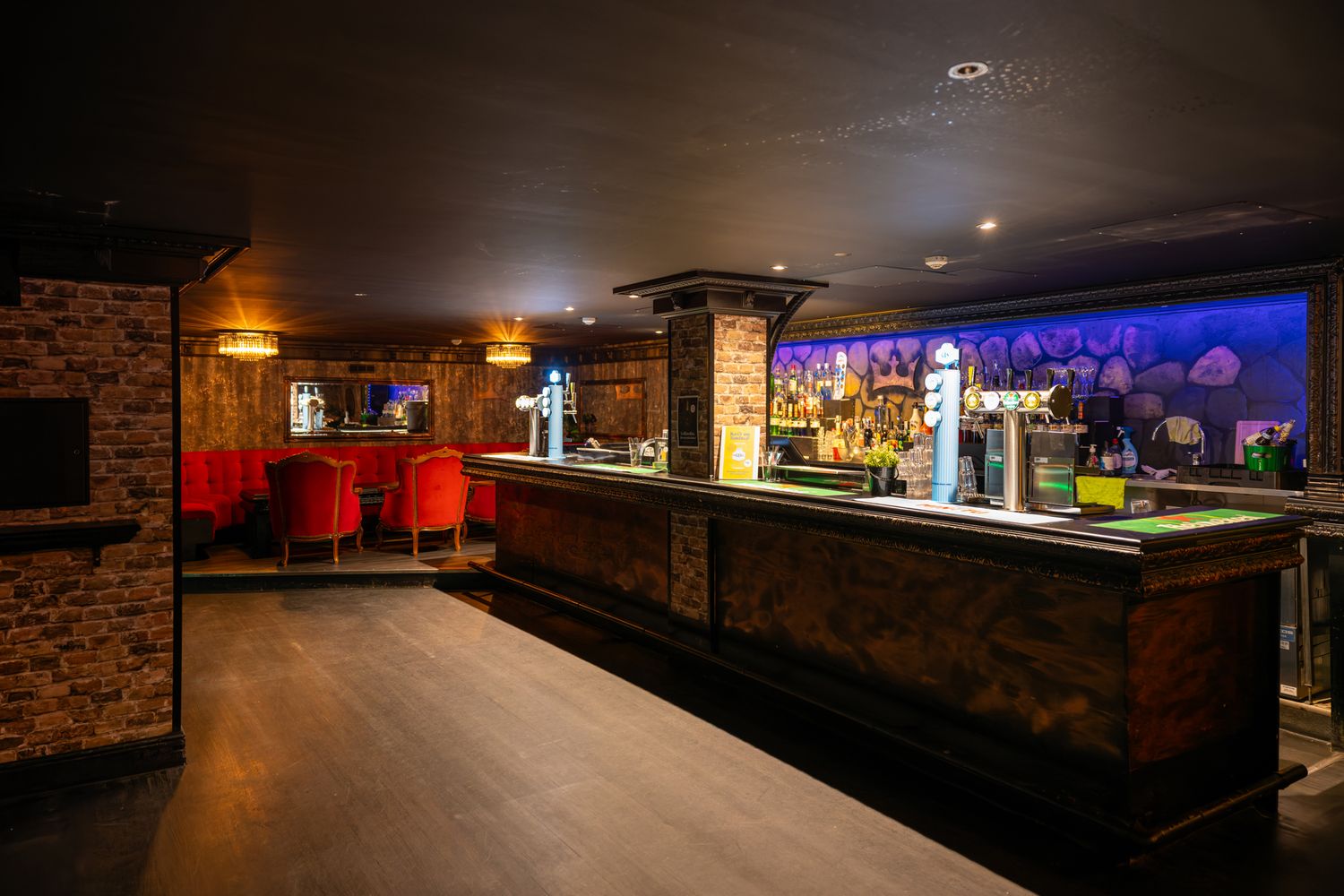Dimly lit bar interior with brick walls, red seating, wood floors, and illuminated back bar with draft taps.