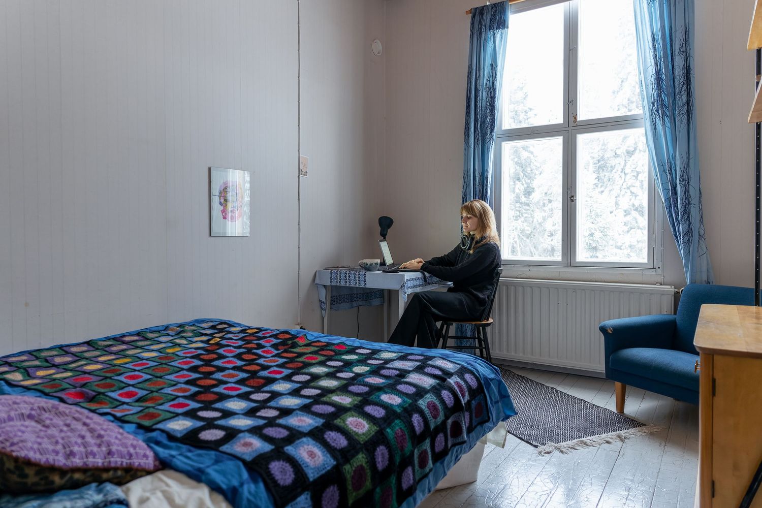 Nainen istuu työpöydän ääressä, ikkunasta avautuu talvinen maisema. Etualalla sänky, jossa sininen virkattu päiväpeite.