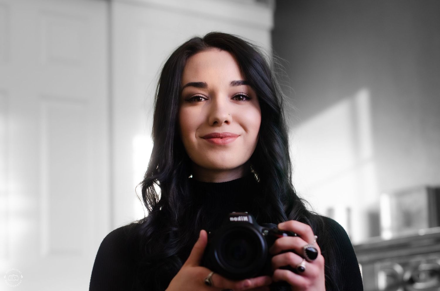 A photographer with dark hair holds a camera for a self-portrait in a light-filled room.