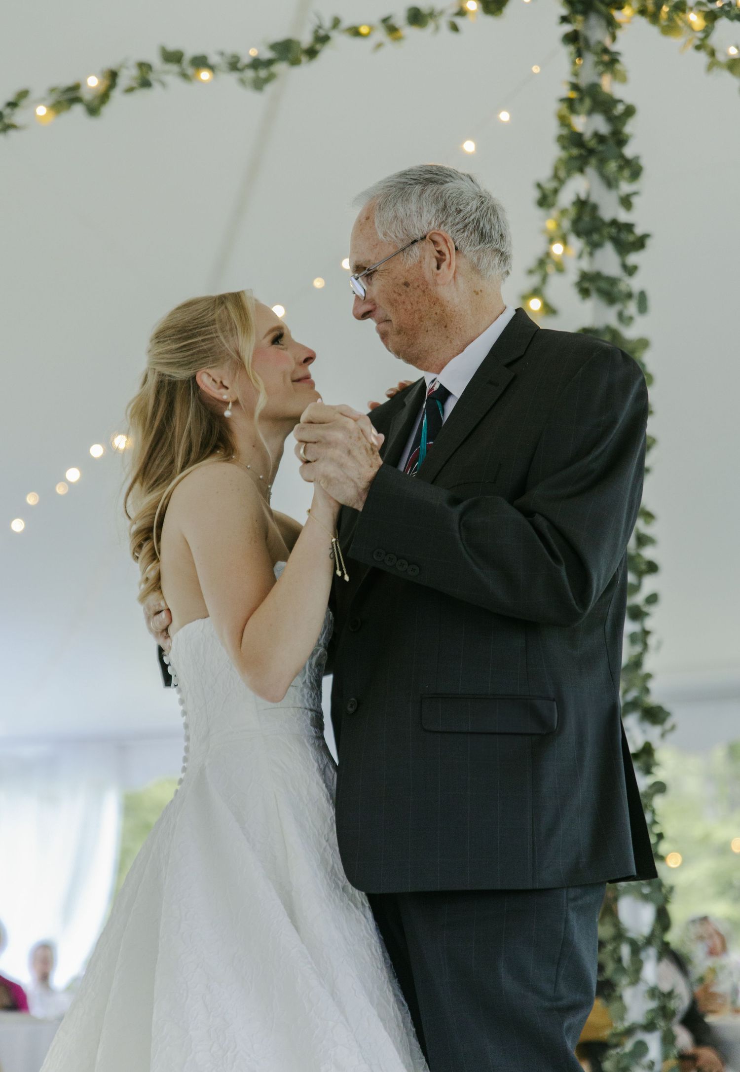 A couple shares a tender dance moment under twinkling lights and green garland at a wedding reception.