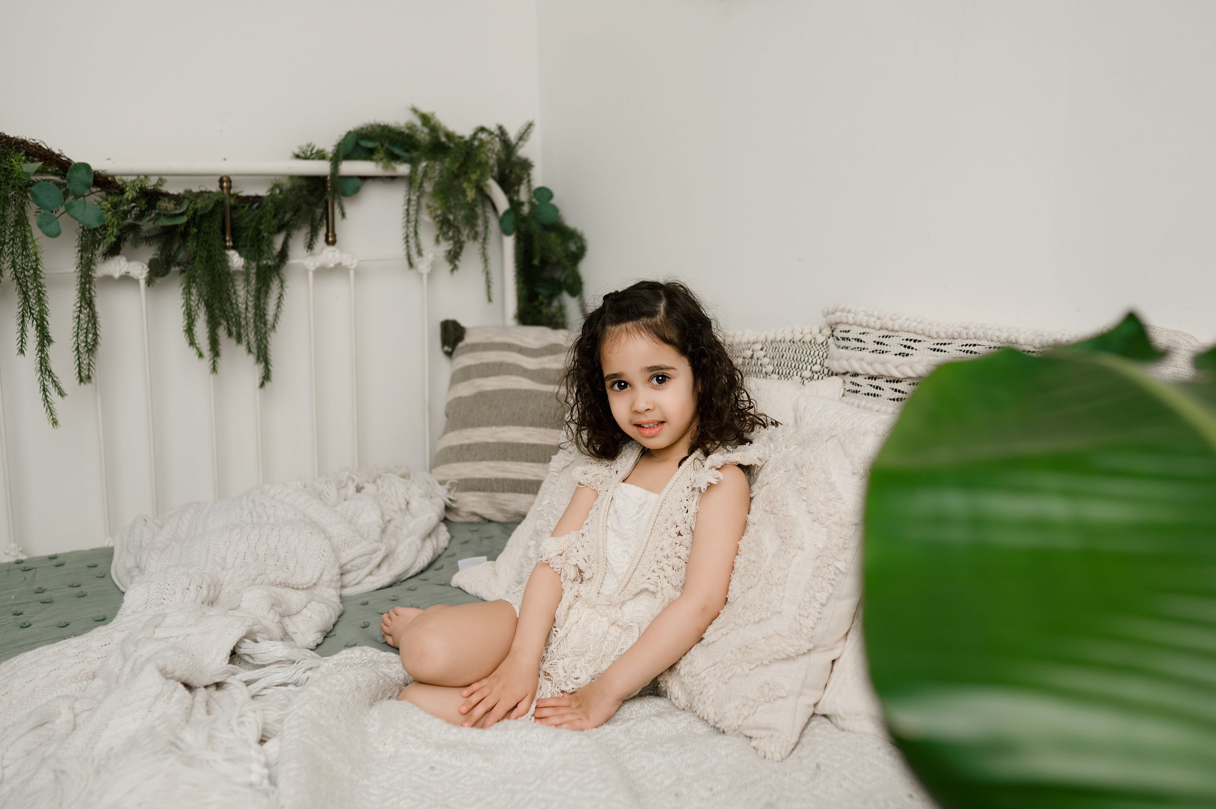 Dreamy boudoir photo series in white lace against soft neutral backdrop with tropical green leaves in foreground.