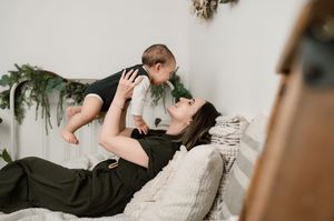 A person in black clothing lies on a cream colored couch while holding and interacting with a baby amid holiday greenery.