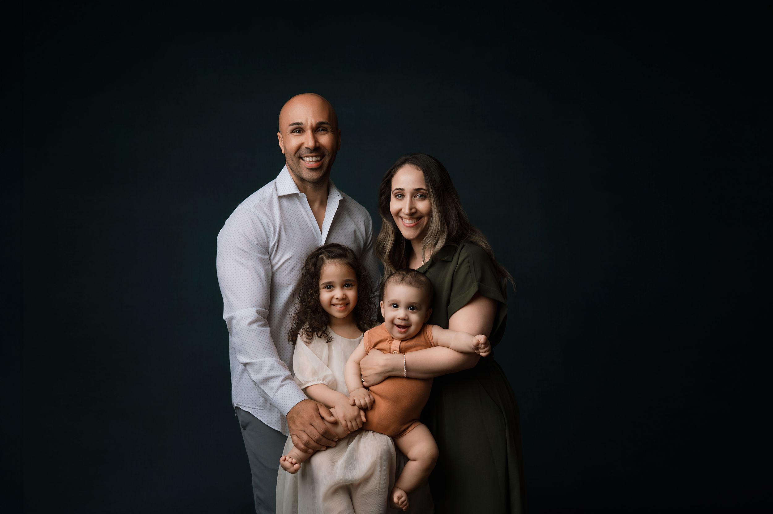 A family of four poses for a portrait against a dark background wearing coordinated casual outfits.
