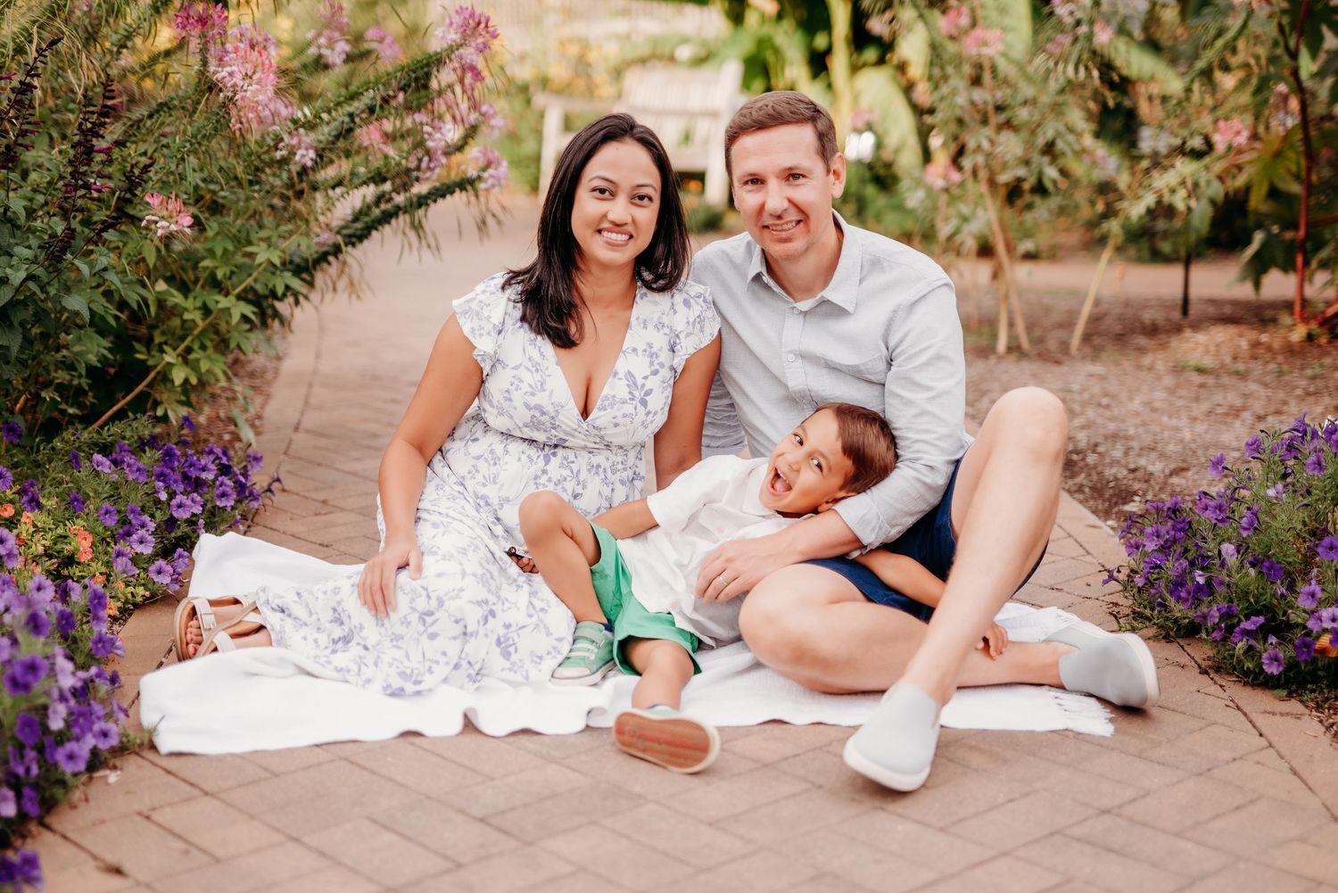 A cheerful family sits together on a blanket in a lush garden with vibrant purple flowers at Horticulture garden at Purdue University