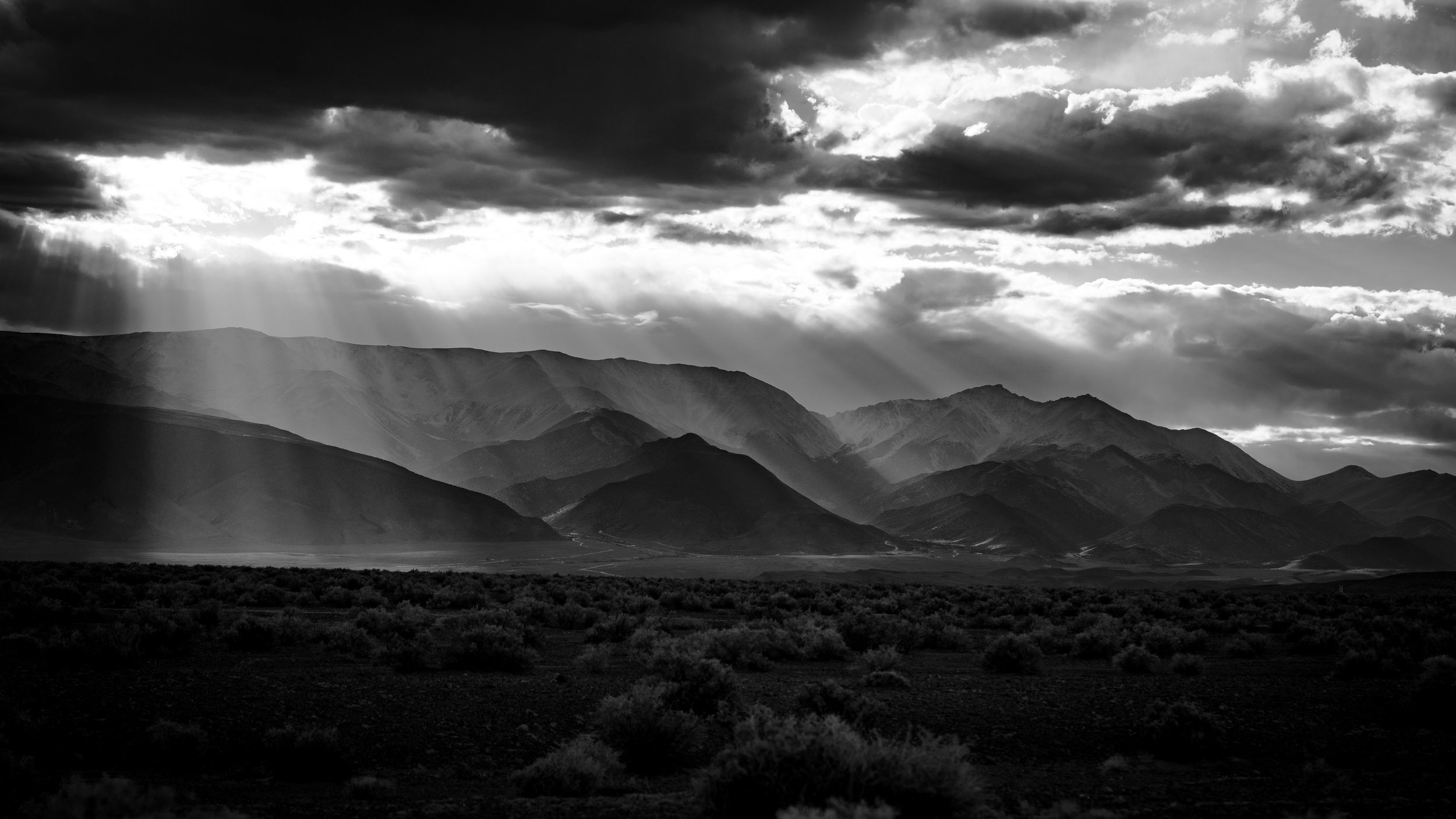 Black and white desert landscape with dramatic sun rays breaking through storm clouds over mountain range.