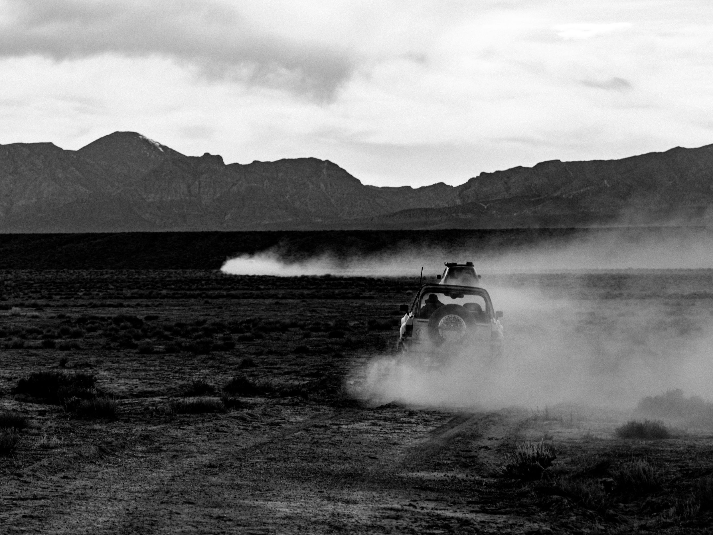 Black and white photo of off-road vehicle speeding across dusty desert with mountain backdrop.