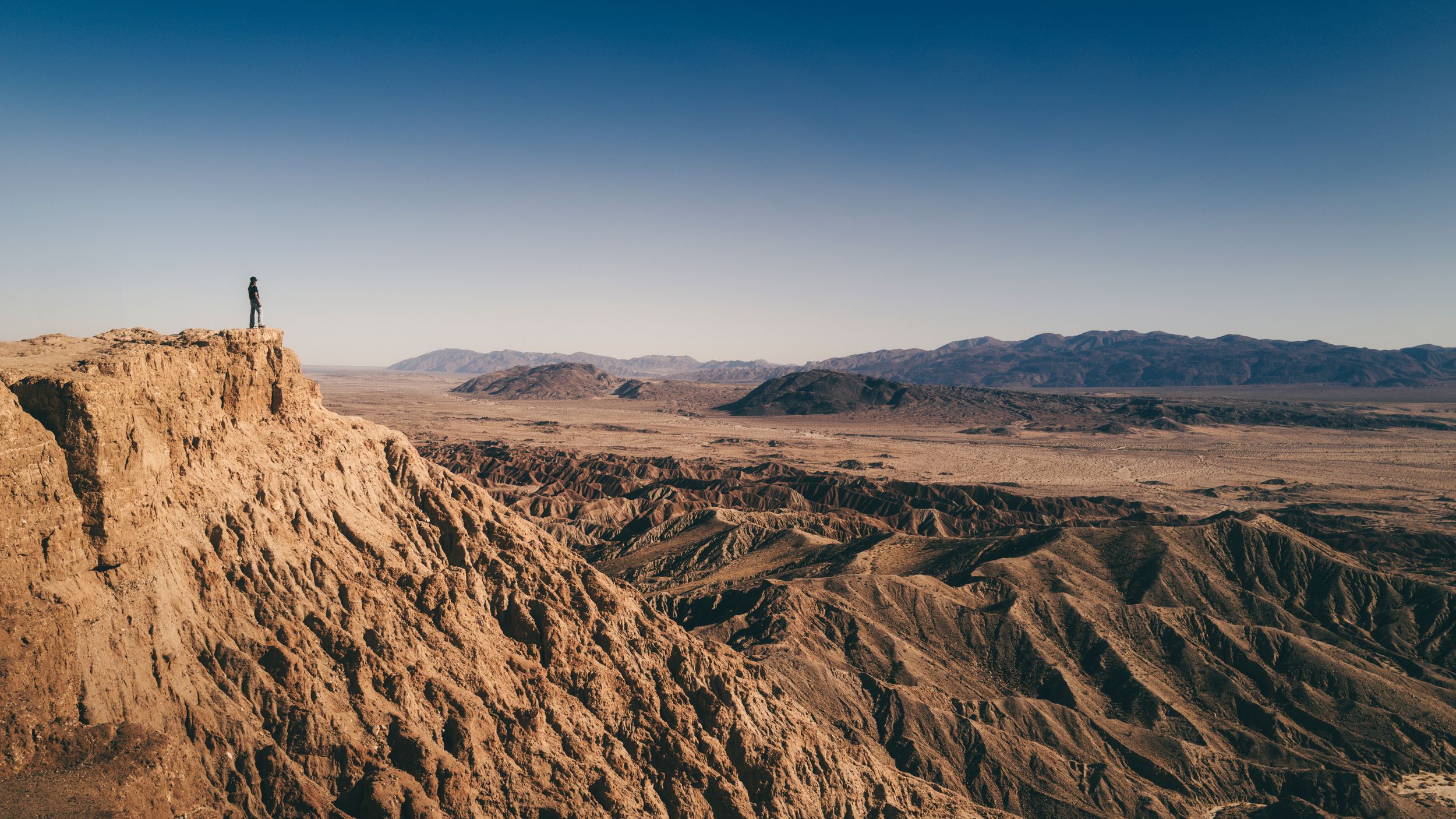 Person standing on rocky desert cliff overlooking vast arid badlands landscape at sunset.