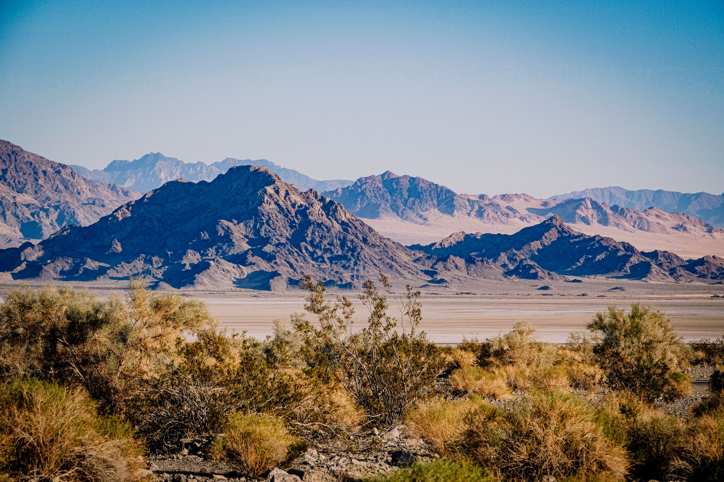 Rocky desert mountain range under clear blue sky with sparse desert vegetation foreground.