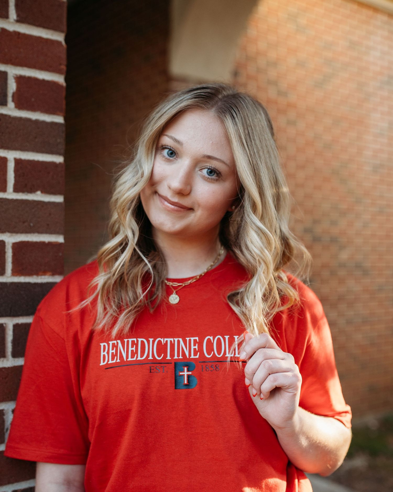 Senior that just graduated high school and is attending Benedictine College wearing her school t-shirt poses against brick wall background.