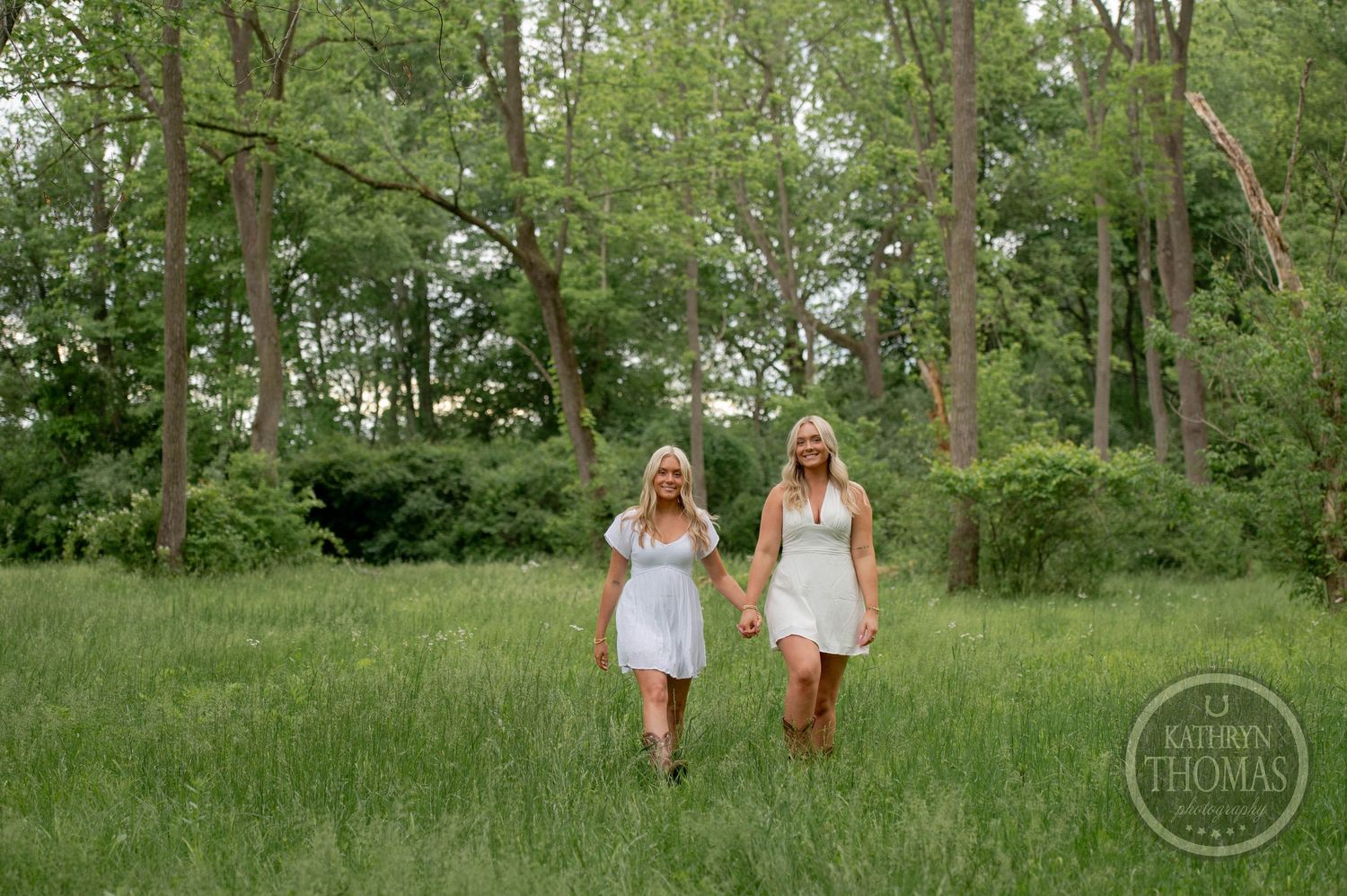 A graduate in white dress and black graduation gown stands in a green woodland meadow at sunset.