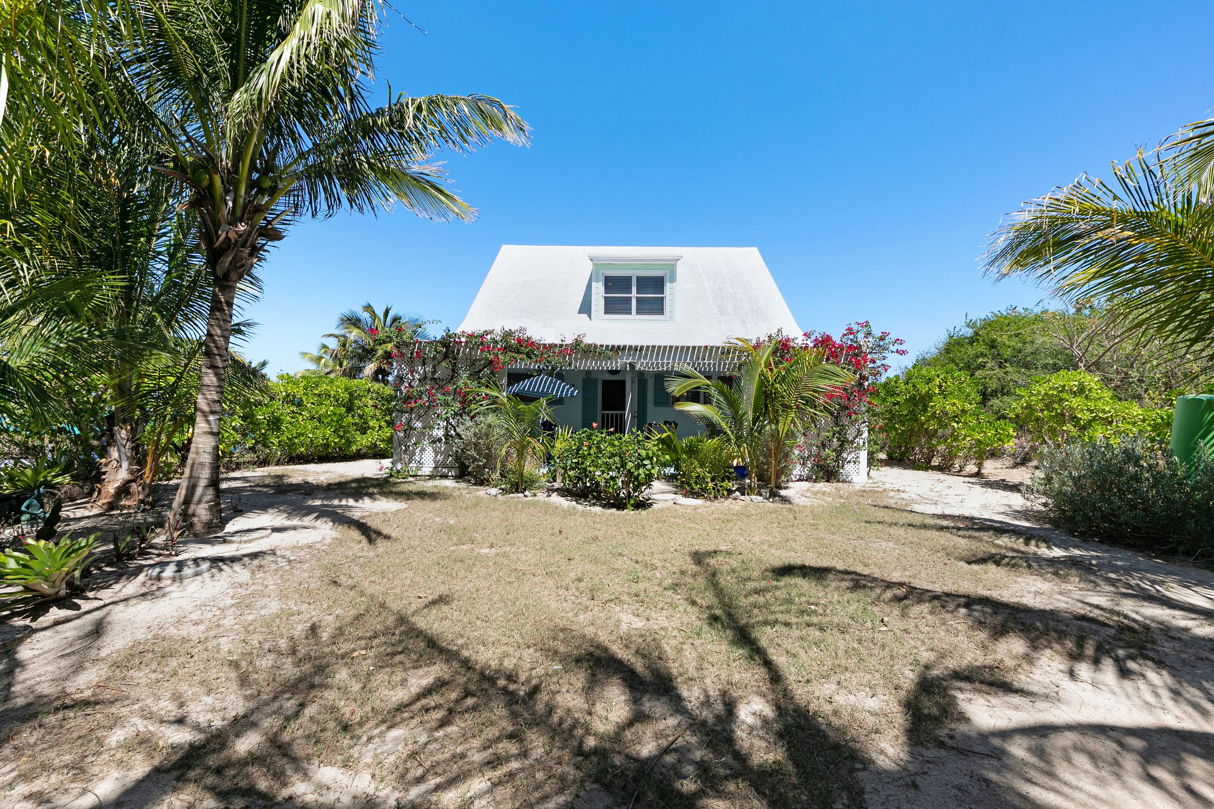 White beach cottage with sand yard surrounded by lush tropical palm trees and landscaping in a sunny setting.